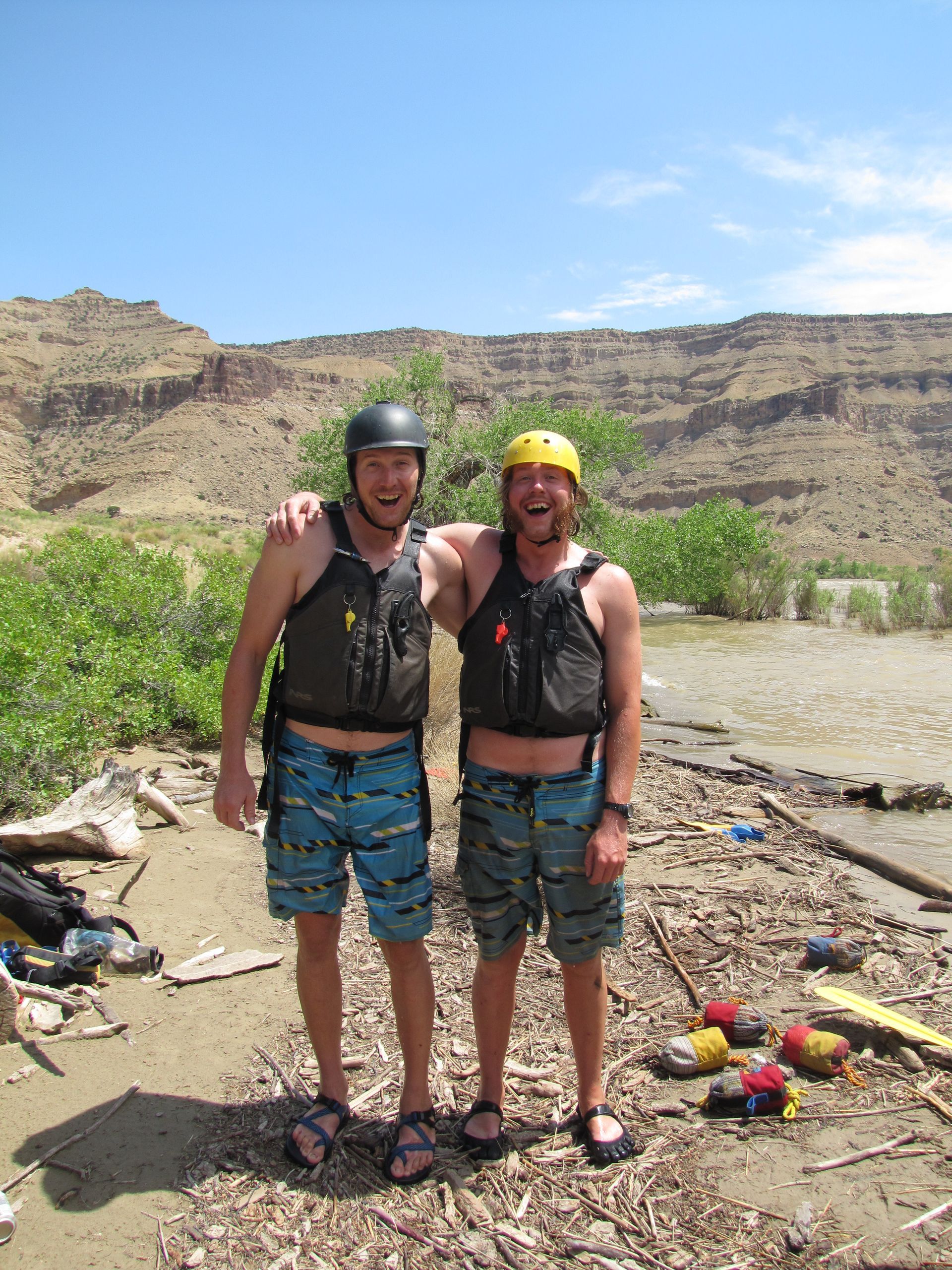 Two men in life vests and helmets pose outdoors; rocky landscape in background.