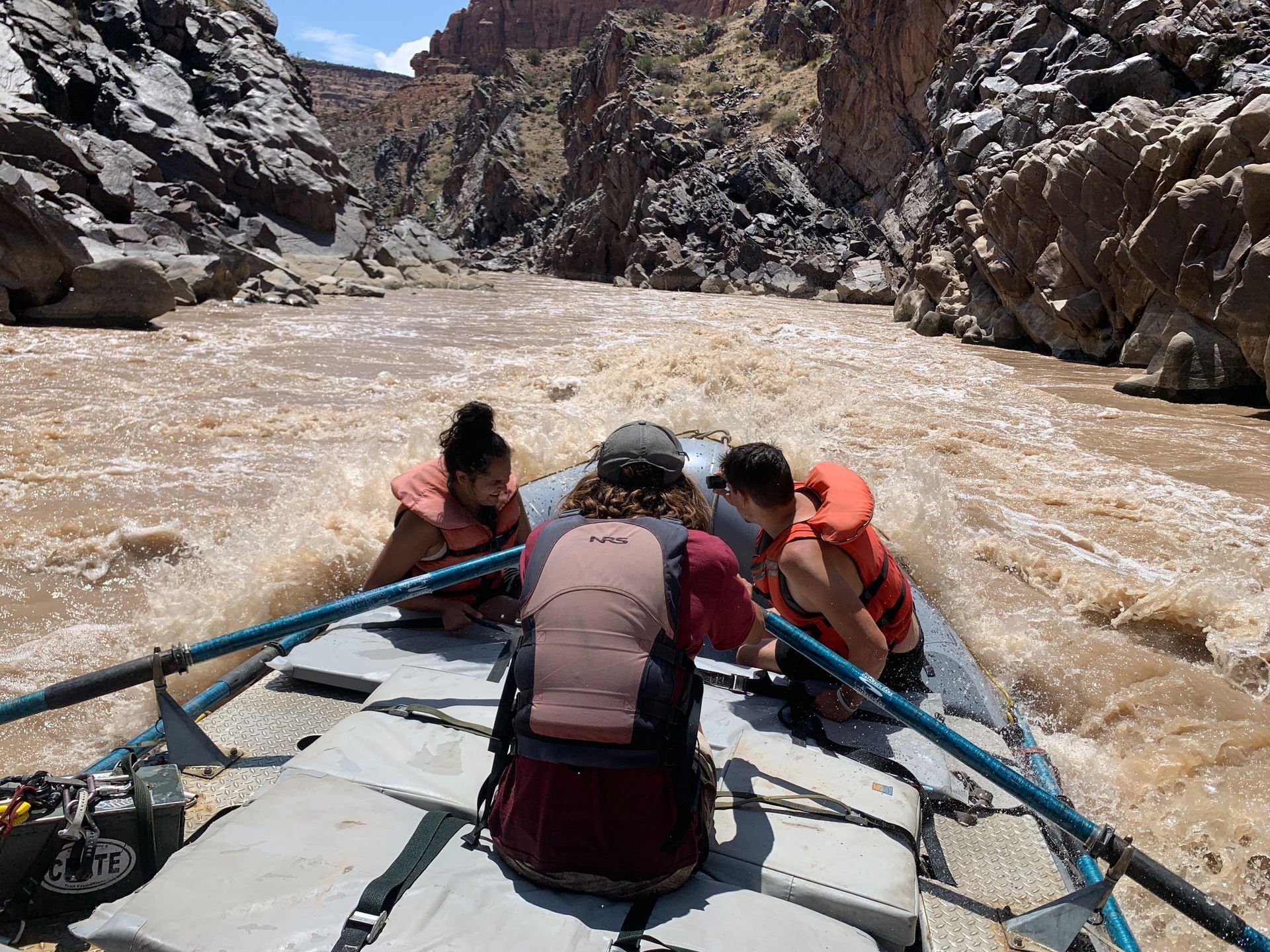 Rafters on a muddy river in a canyon. One person rows, two in orange life vests brace themselves.