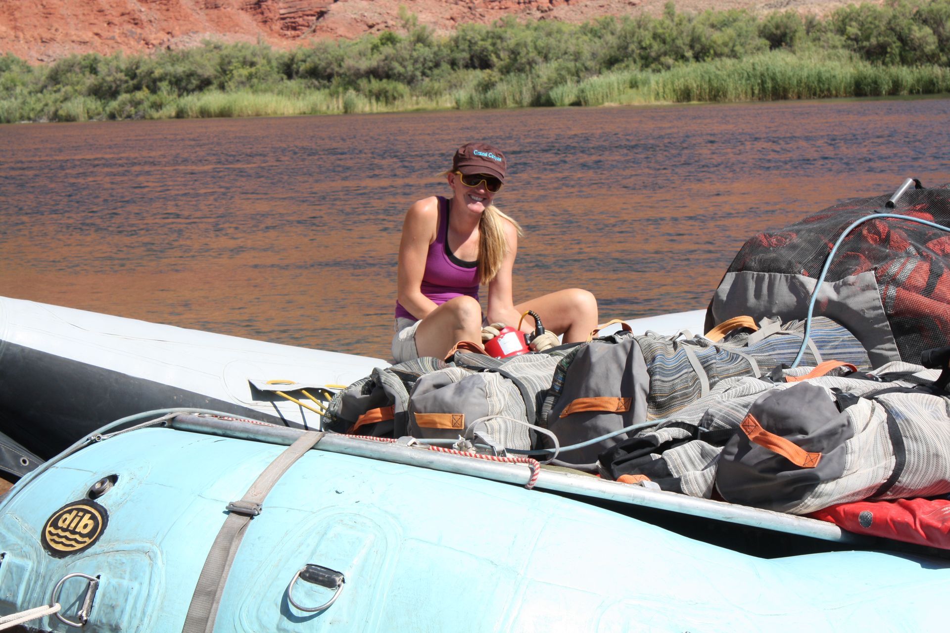 Woman on a raft, smiling. Raft is loaded with gear on a river with red rock in the background.