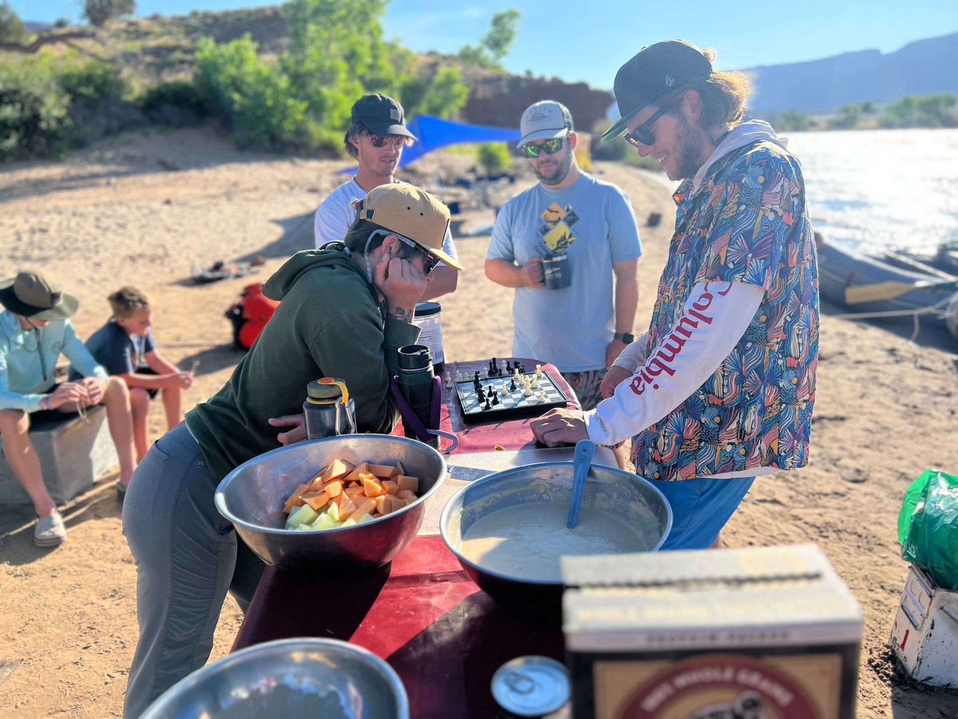 People preparing food at a river bank. Bowls of food on a table. Others watch.