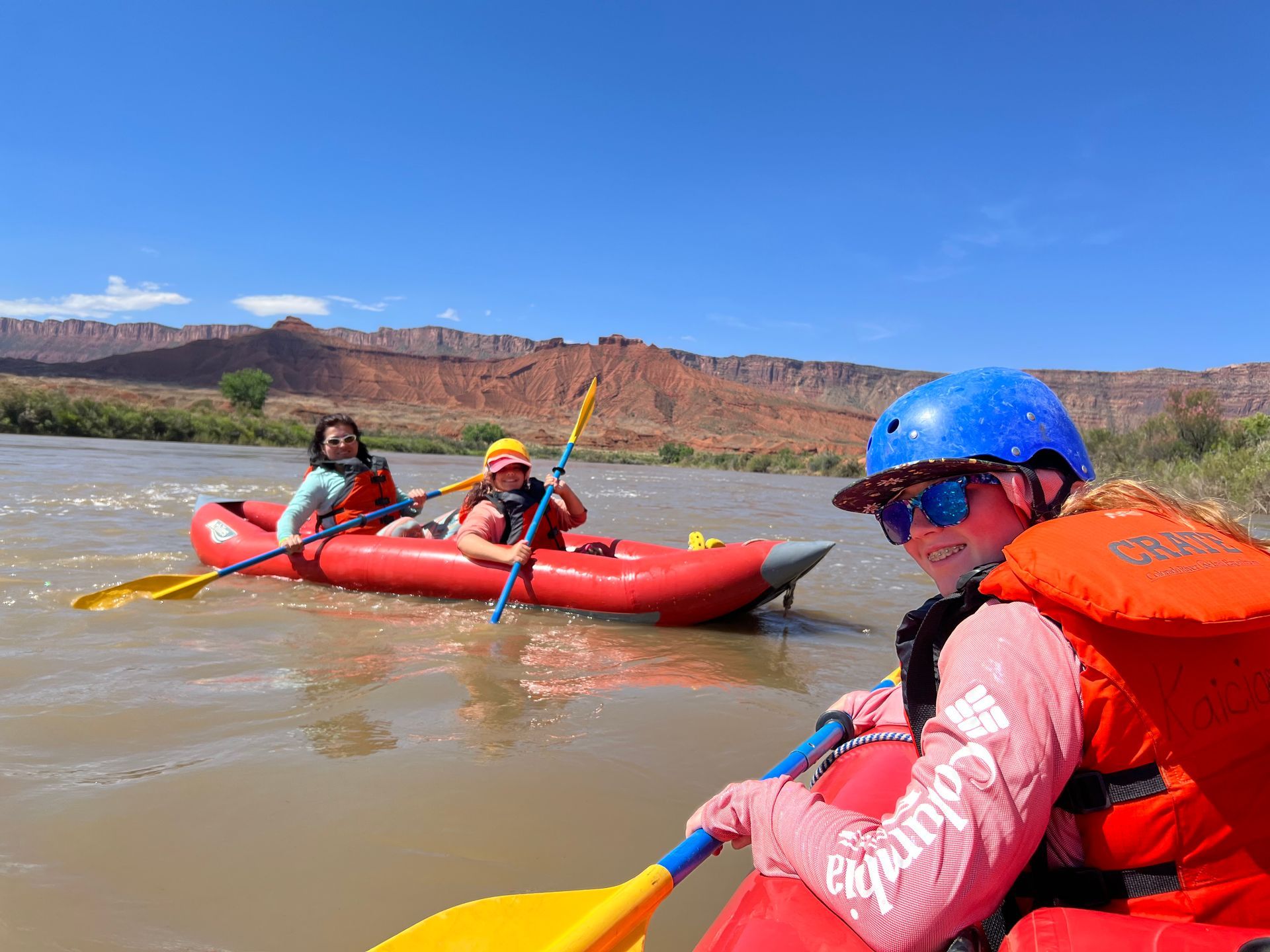 People in a red raft paddling on a river, blue sky, red rock mountains in the background.