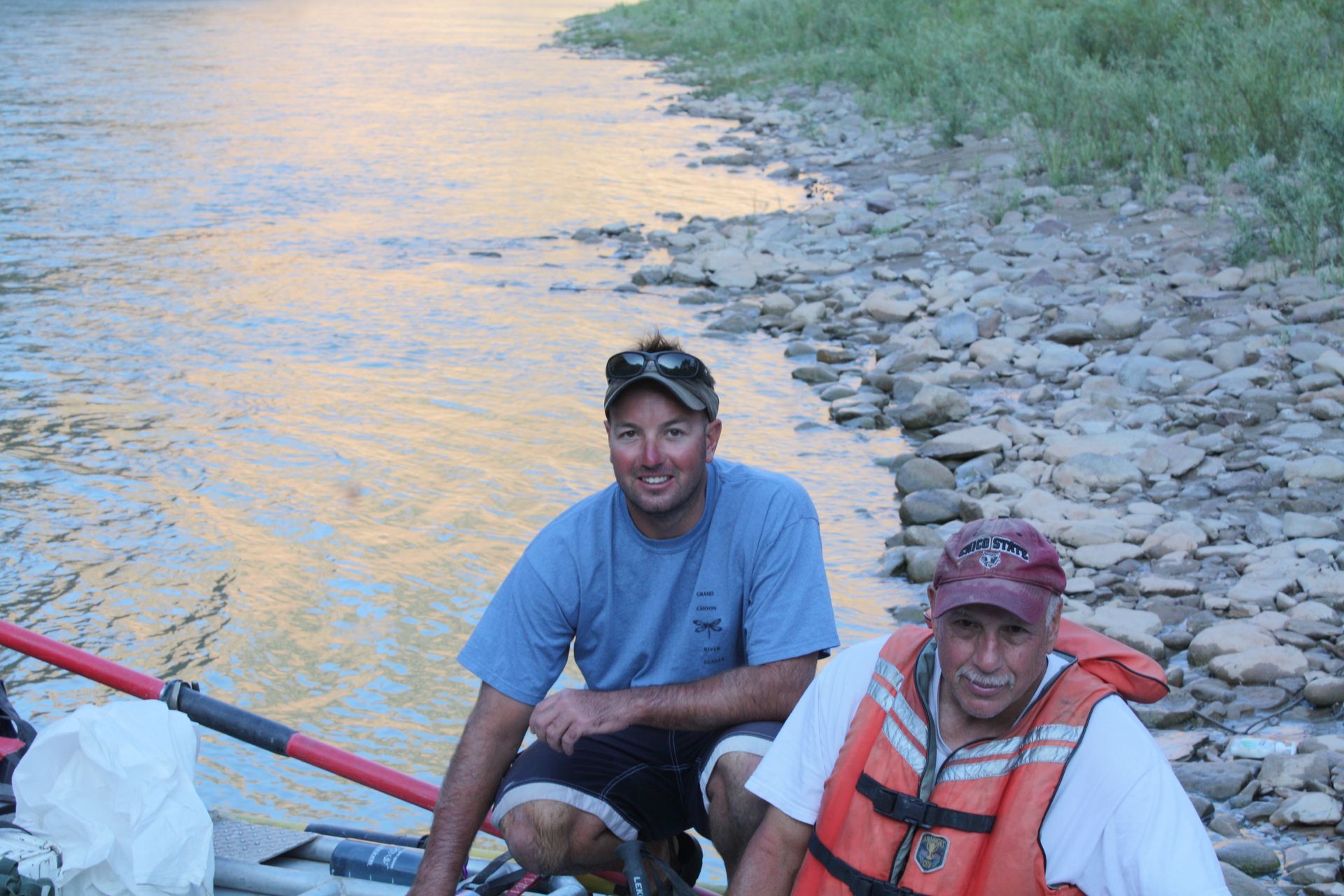 Two men in a raft on a river. One kneels, one wears a life vest, rocky shore, calm water with sunset reflections.