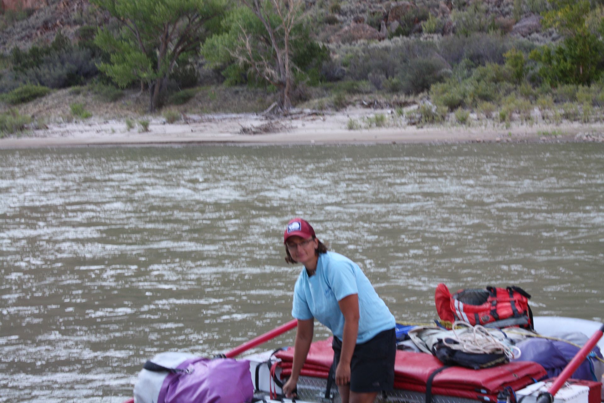 Person on a raft smiles at the camera, with gear visible. River and shoreline in background.