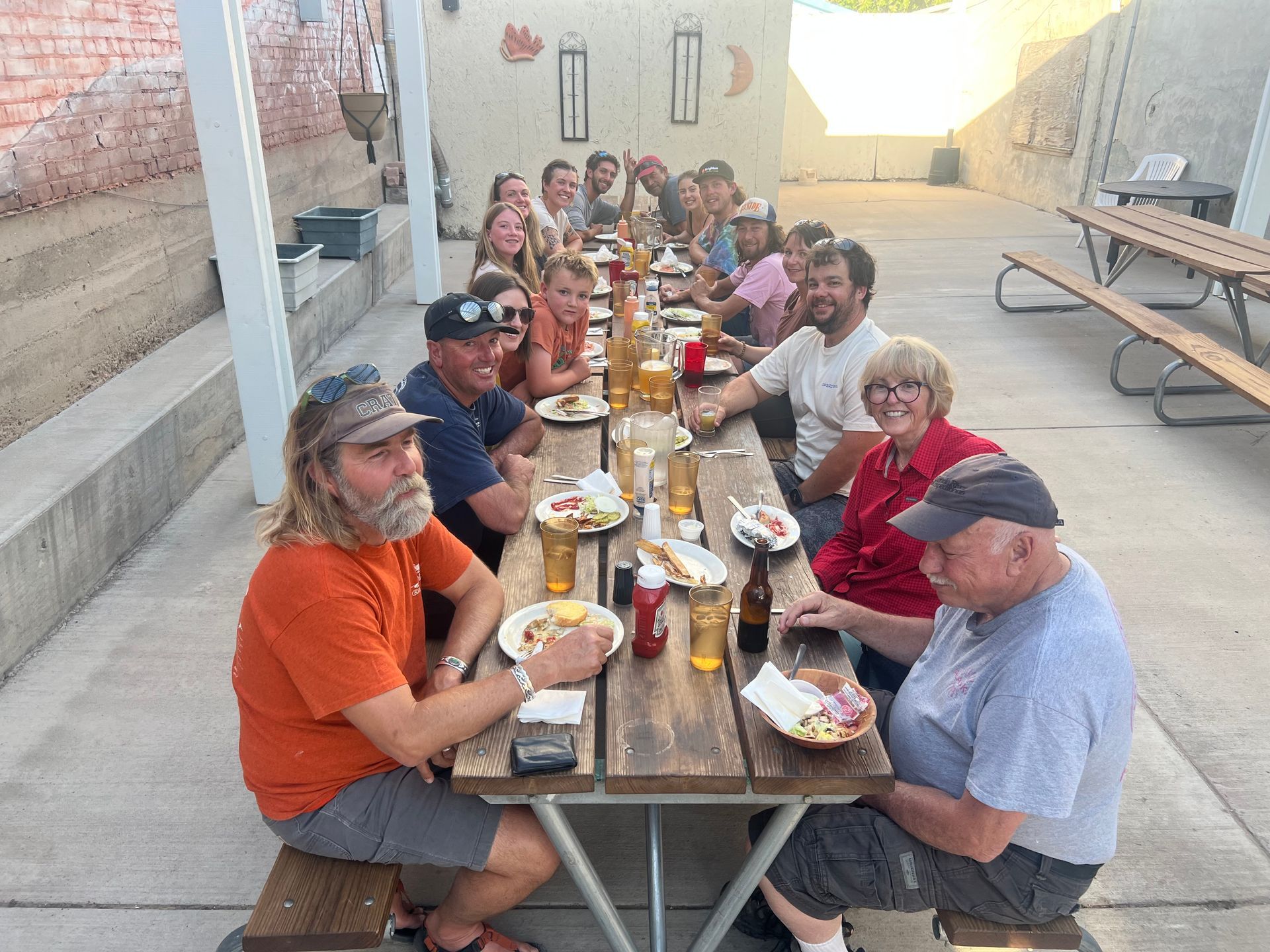 People seated at a long outdoor table, eating and drinking. Brick wall in background.