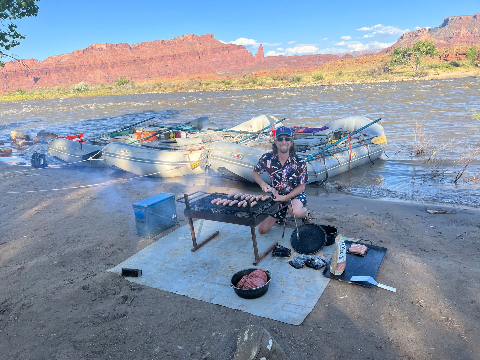 Person grilling food near rafts on a riverbank, red rock backdrop, sunny day.