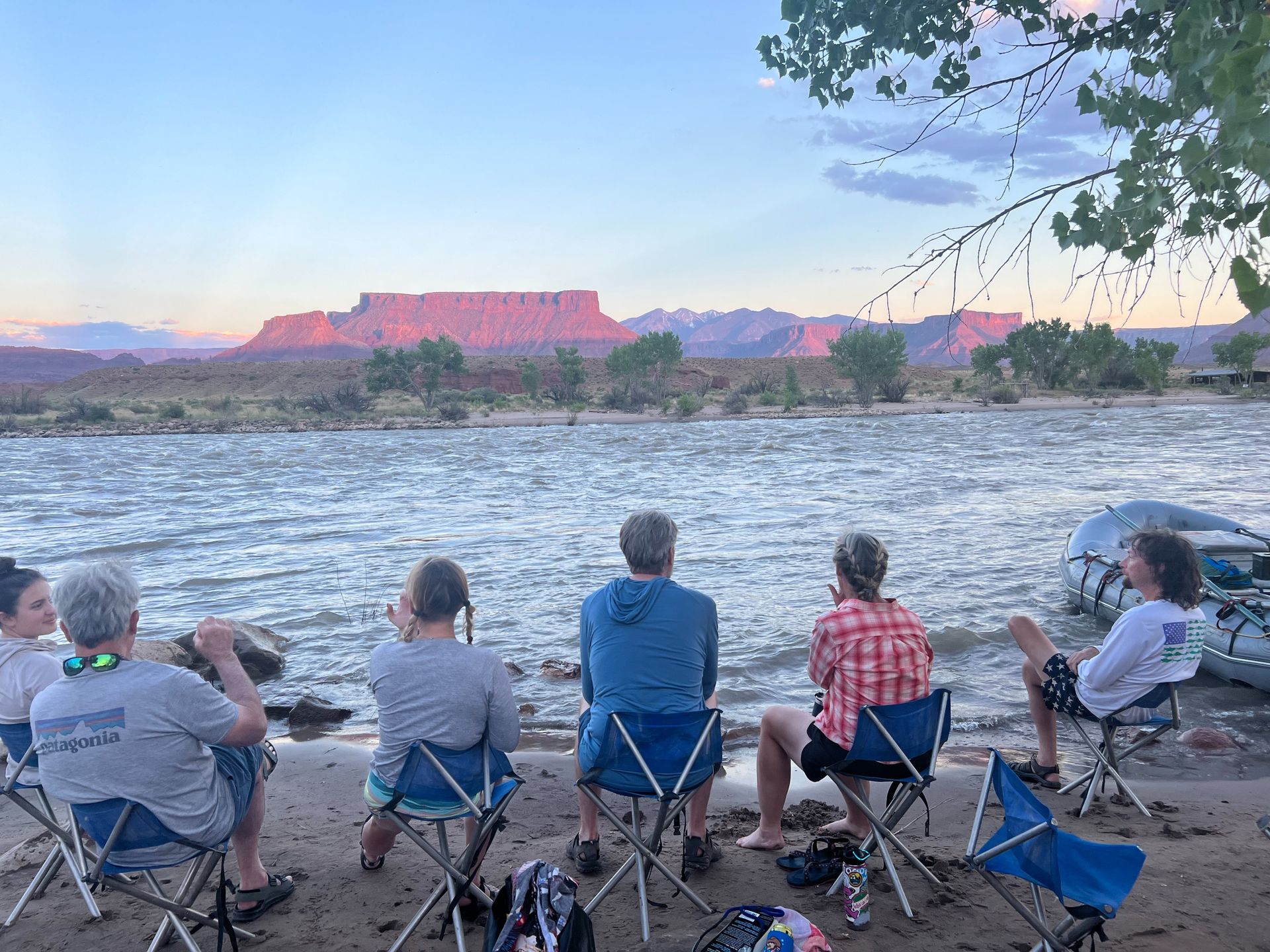 People in camping chairs on a riverbank, gazing at a distant red rock mountain at sunset.