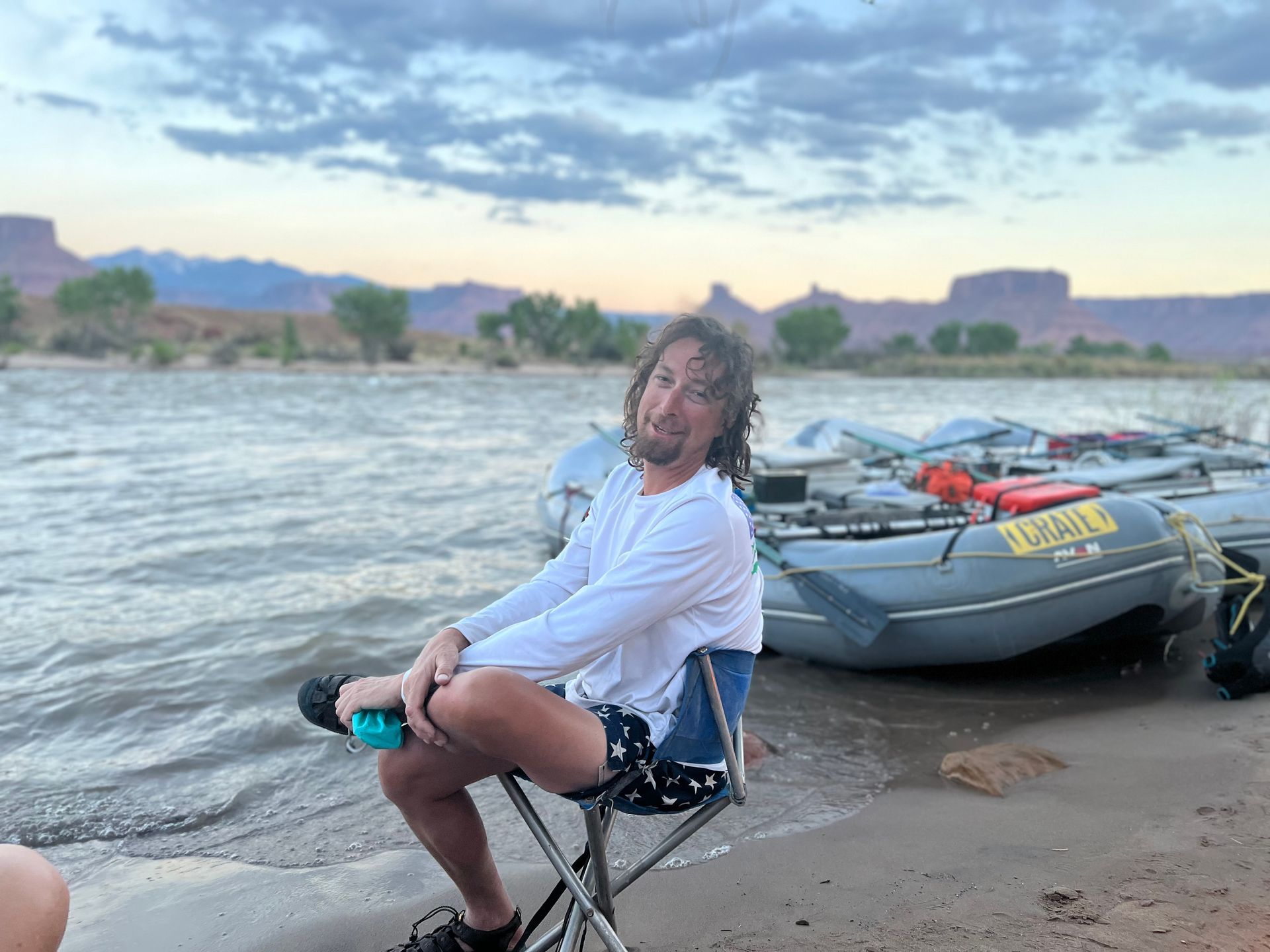 Man sitting on a folding chair on a beach by a river, with rafts in the background, dusk setting.