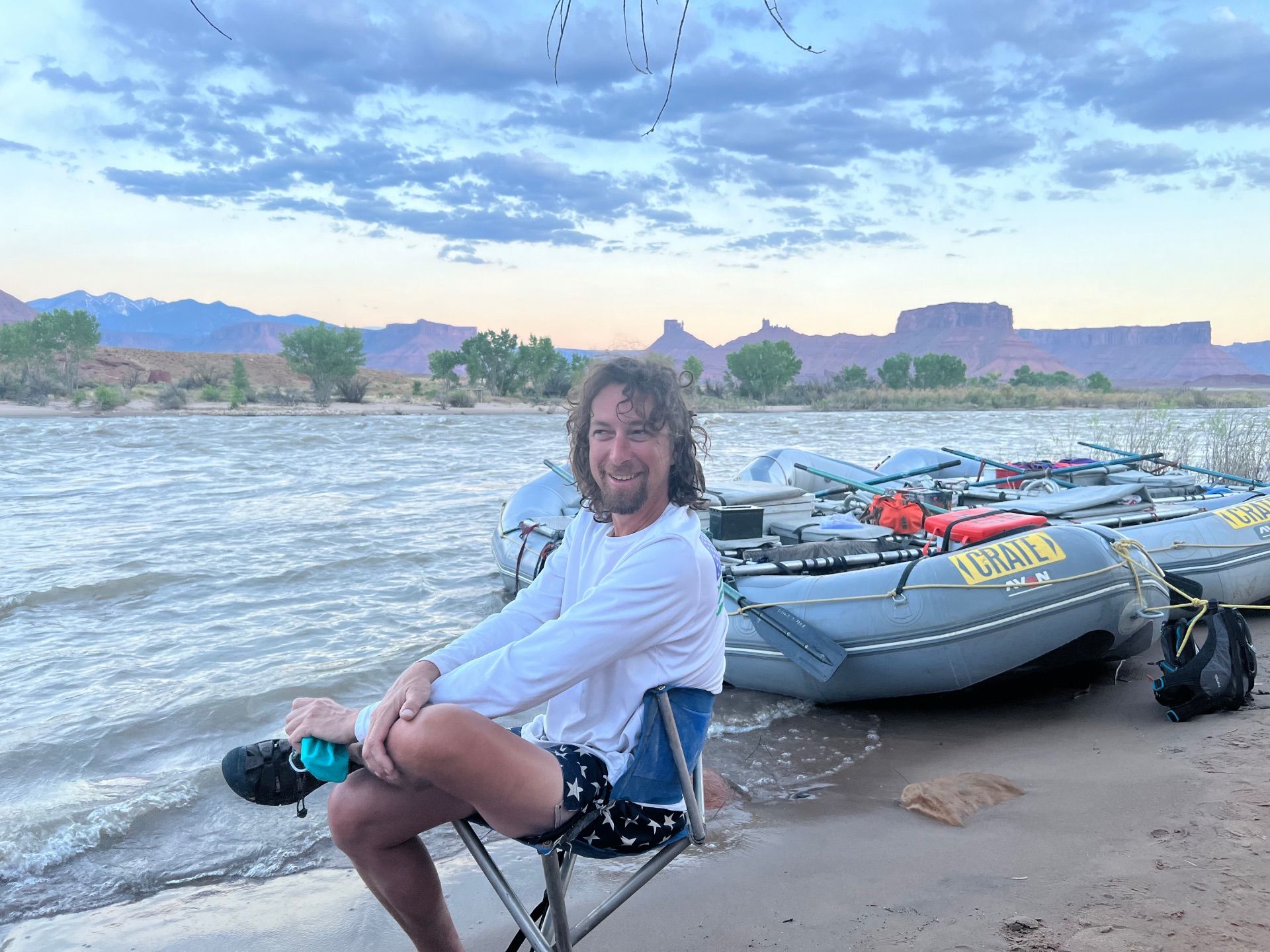Man sitting in a chair by a river with rafts, reddish mountains, and cloudy sky in background.