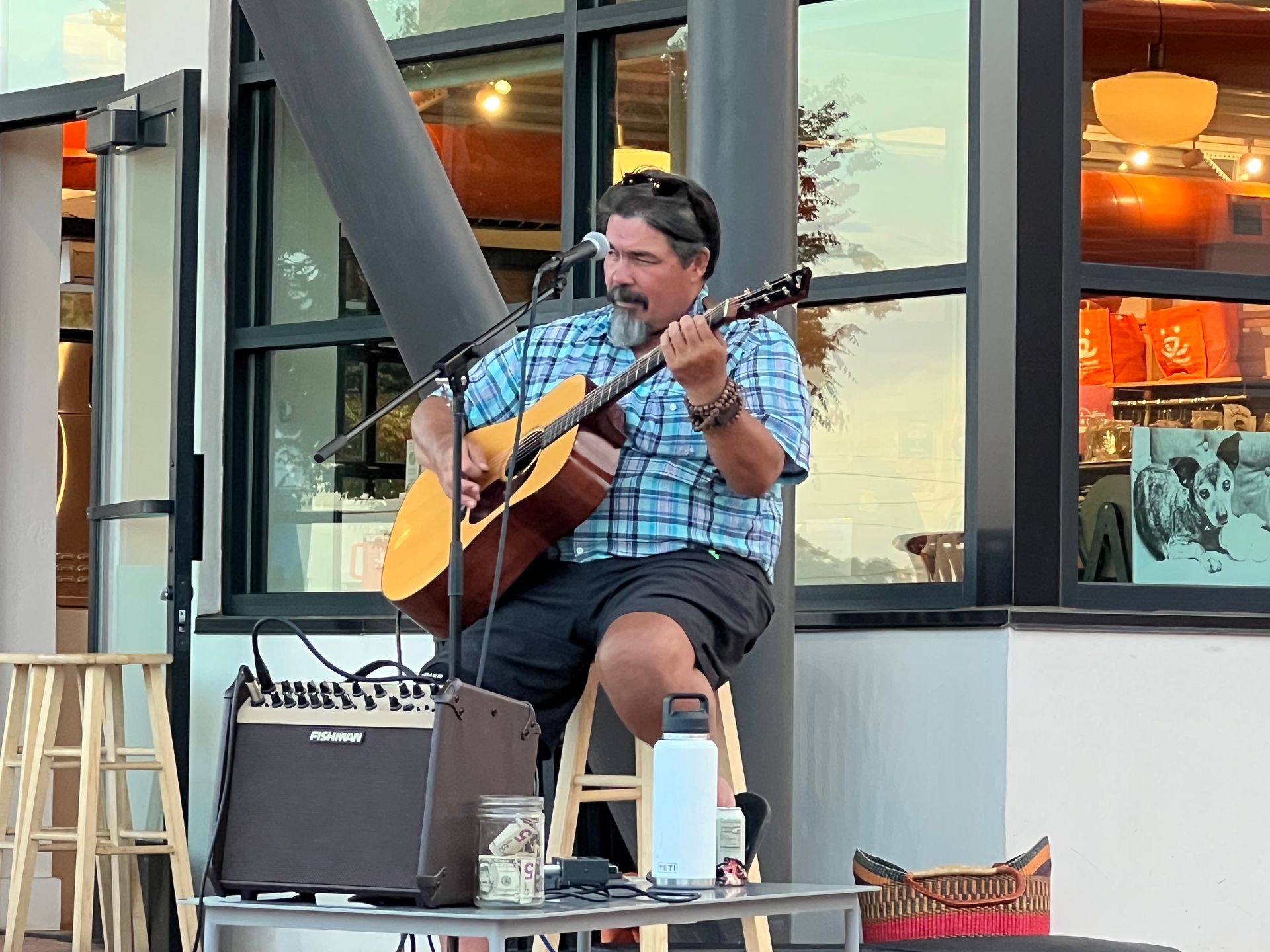 Man plays acoustic guitar outdoors, sitting on a stool. Amplifier, jar for tips, and water bottle nearby.