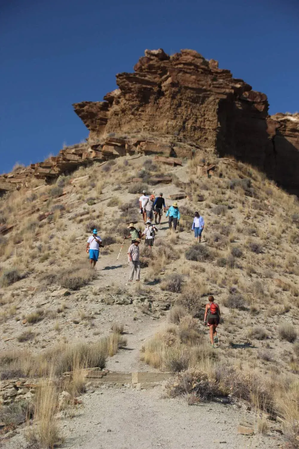 Hikers ascend a dusty, sloped trail towards a rocky outcropping under a clear blue sky.