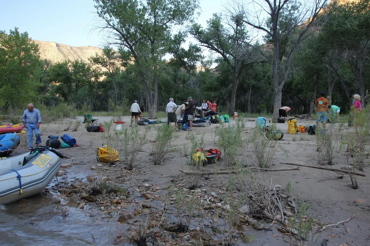 Camp scene with people and gear near a river, with rafts on the shore and trees in the background.