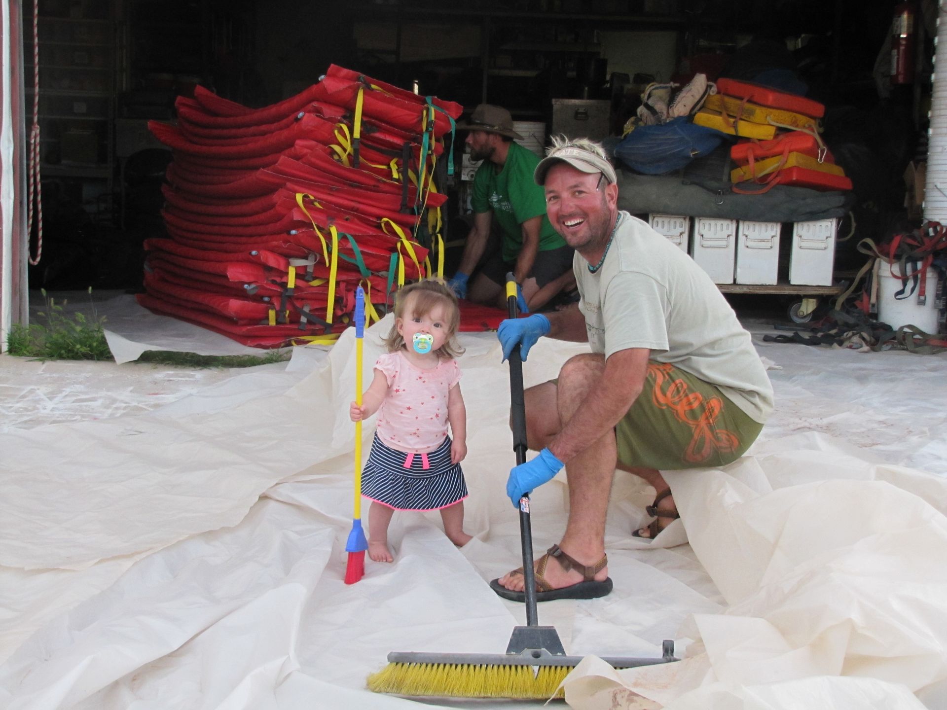 Man and toddler with pacifier on tarp, cleaning with brooms, red rafts in background.