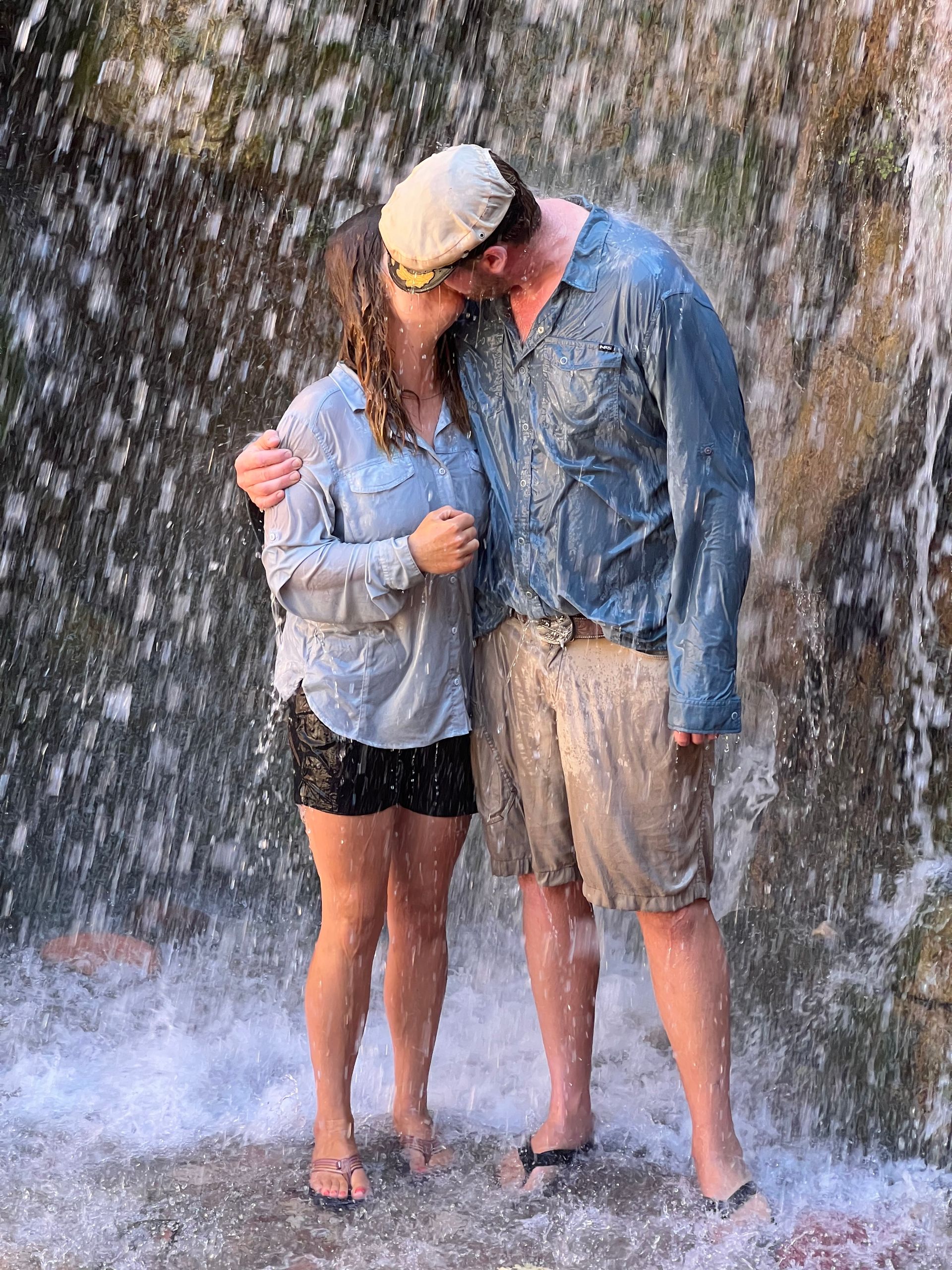 Couple kissing under waterfall; both soaked, wearing casual clothes.