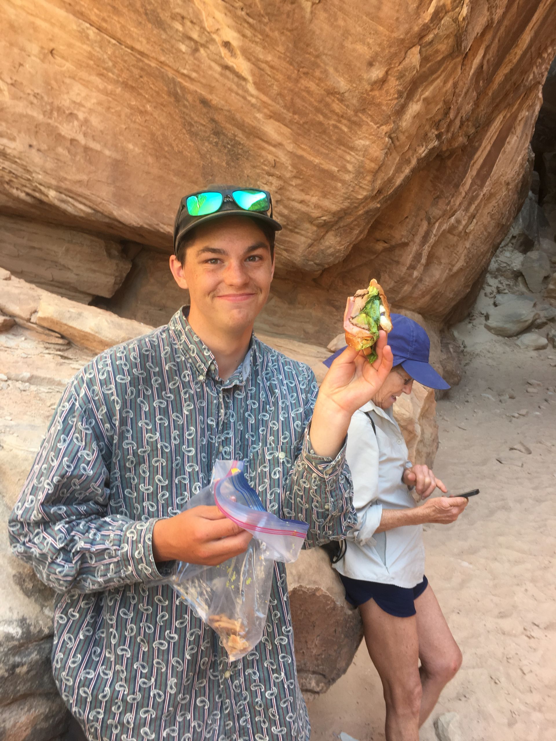 Man holding a sandwich and a bag of food smiling in front of a rock formation; person in background on phone.