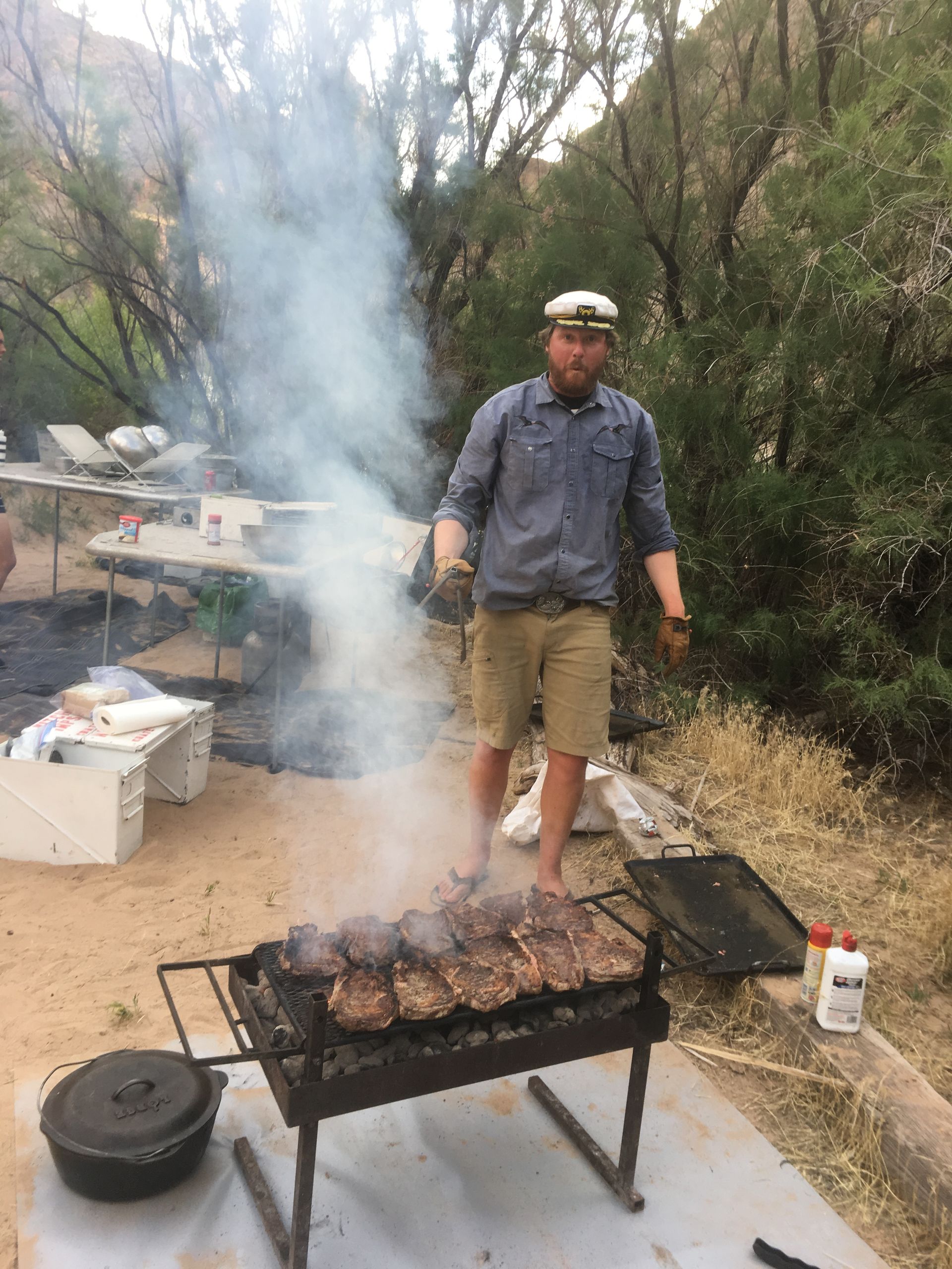 Man grilling ribs outdoors, surrounded by smoke. He's wearing a hat, shirt, shorts.
