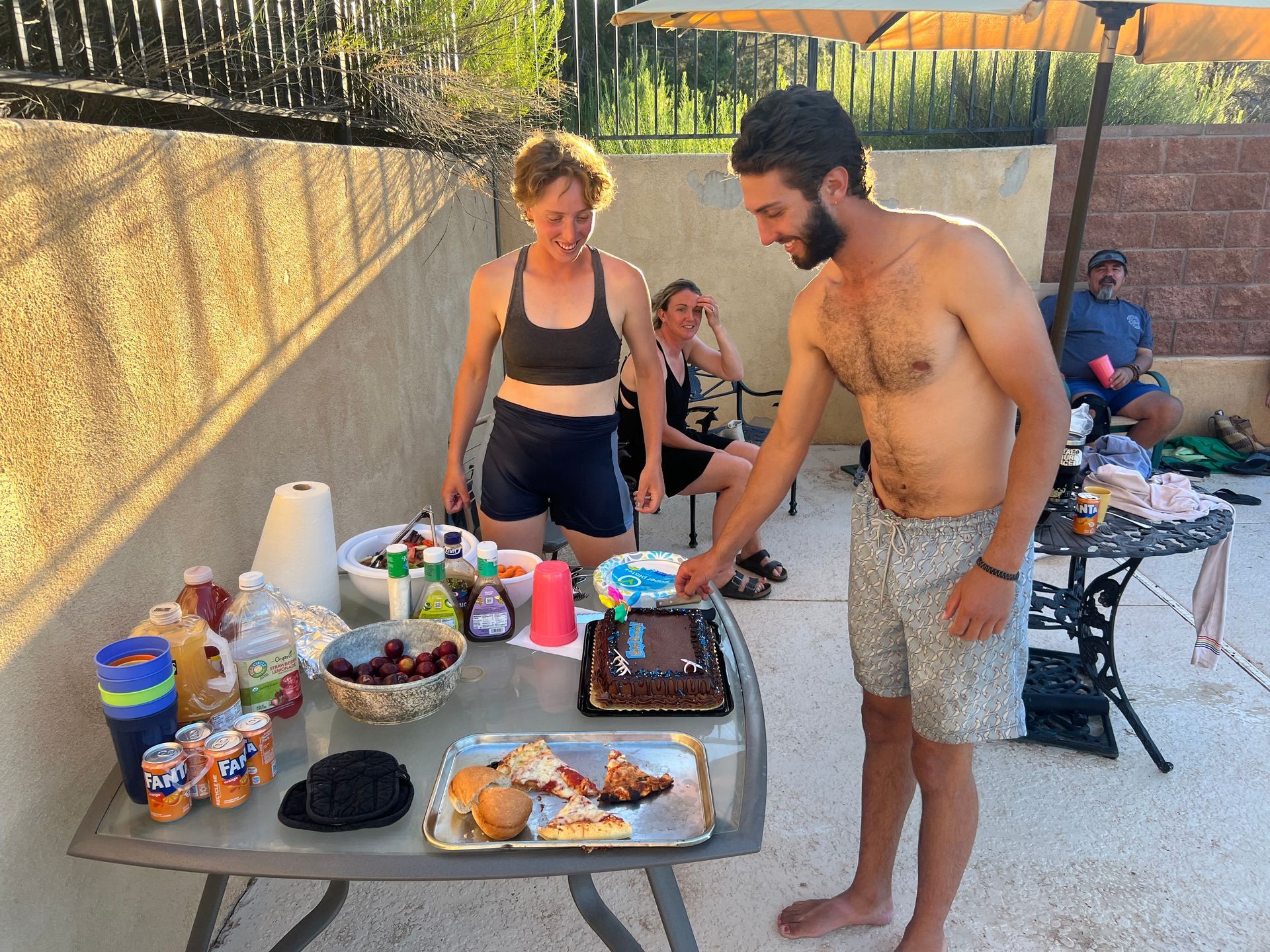 People at outdoor party. Man cuts cake; two women watch. Food/drinks on table. Sunny, patio setting.