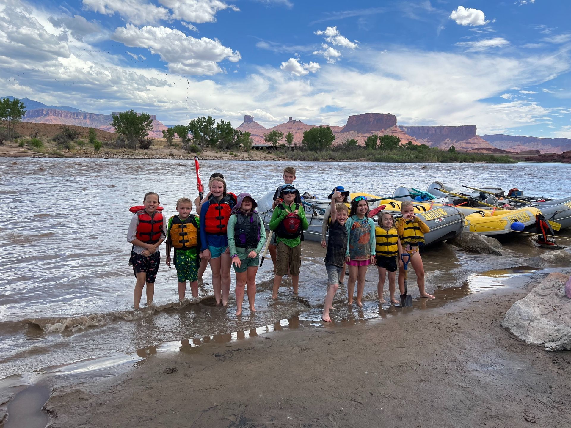 Group of children in life vests standing near rafts on a riverbank, mountains in the background.