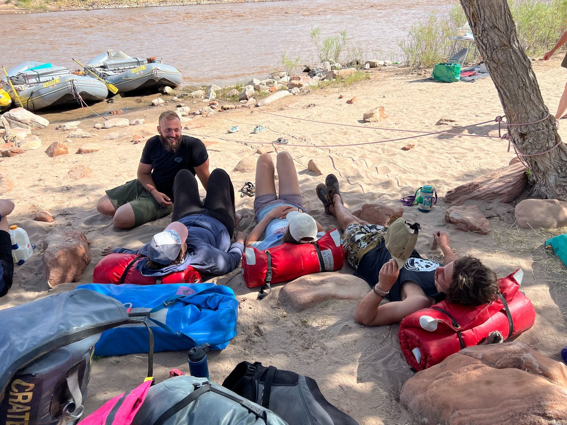 People resting on sand near a river, with red dry bags, a blue duffel, and rafts in the background.