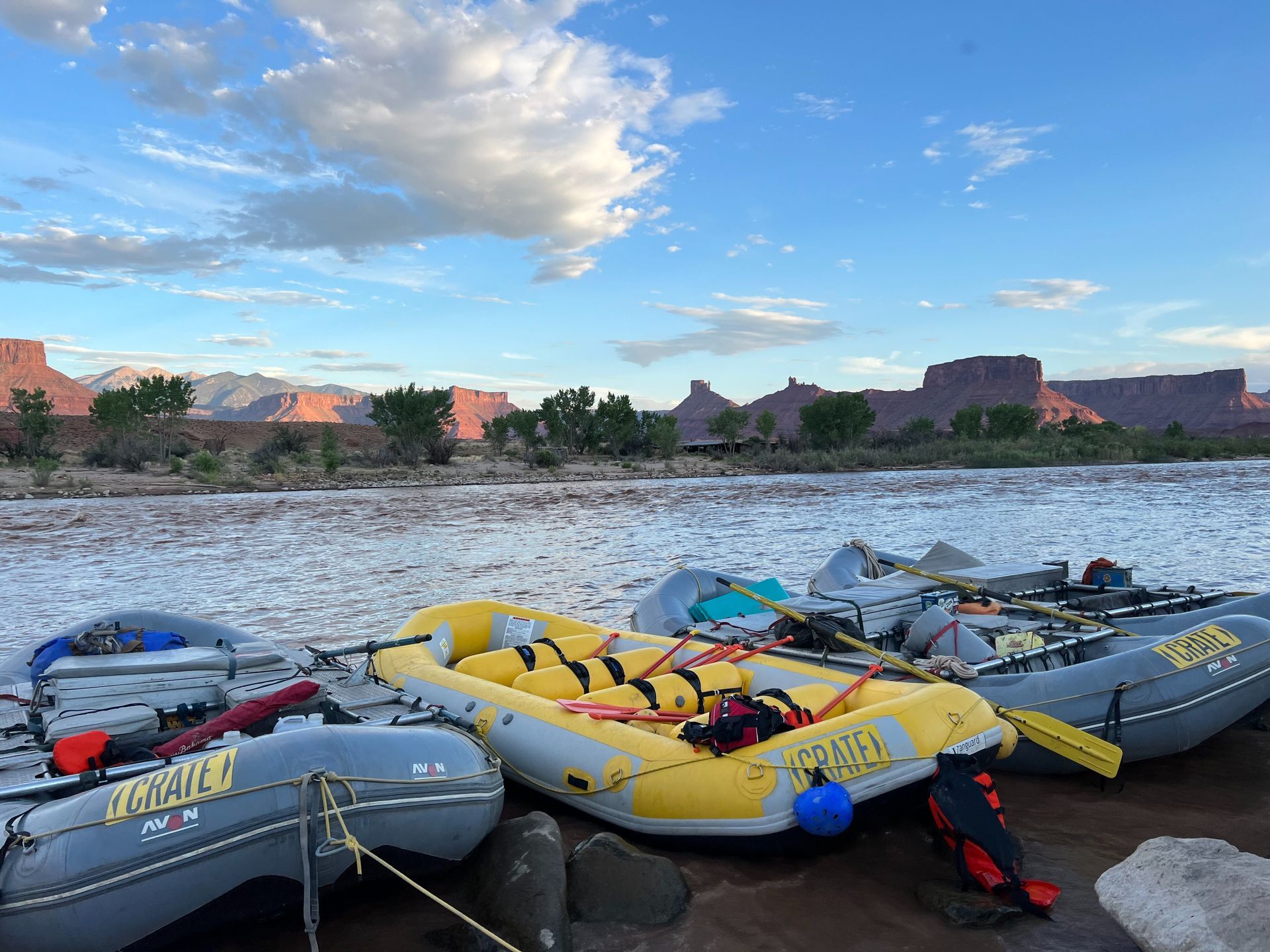 Rafting boats on a river bank with red rock formations in the background under a blue cloudy sky.