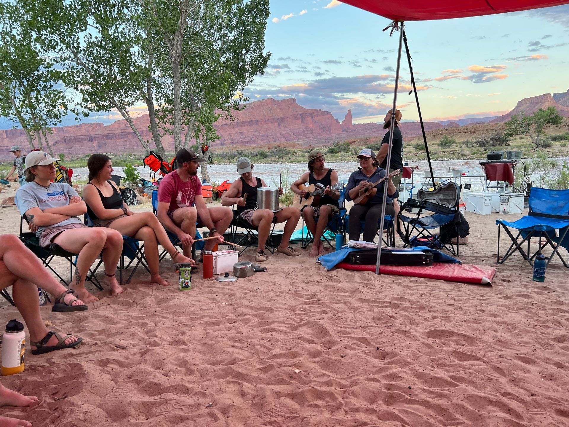 People on a sandy riverbank, gathered for music. Red-rock cliffs and a river in the background, under a canopy.
