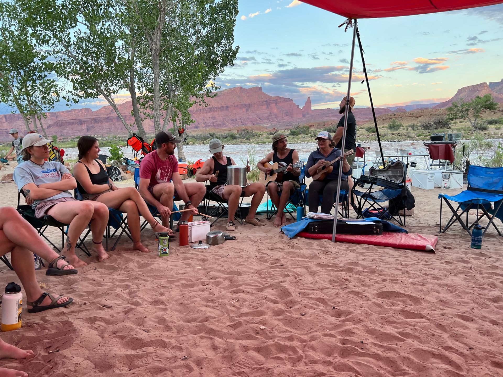 Group on a sandy bank playing music. Red rock backdrop, under a canopy. Evening sky, water nearby.