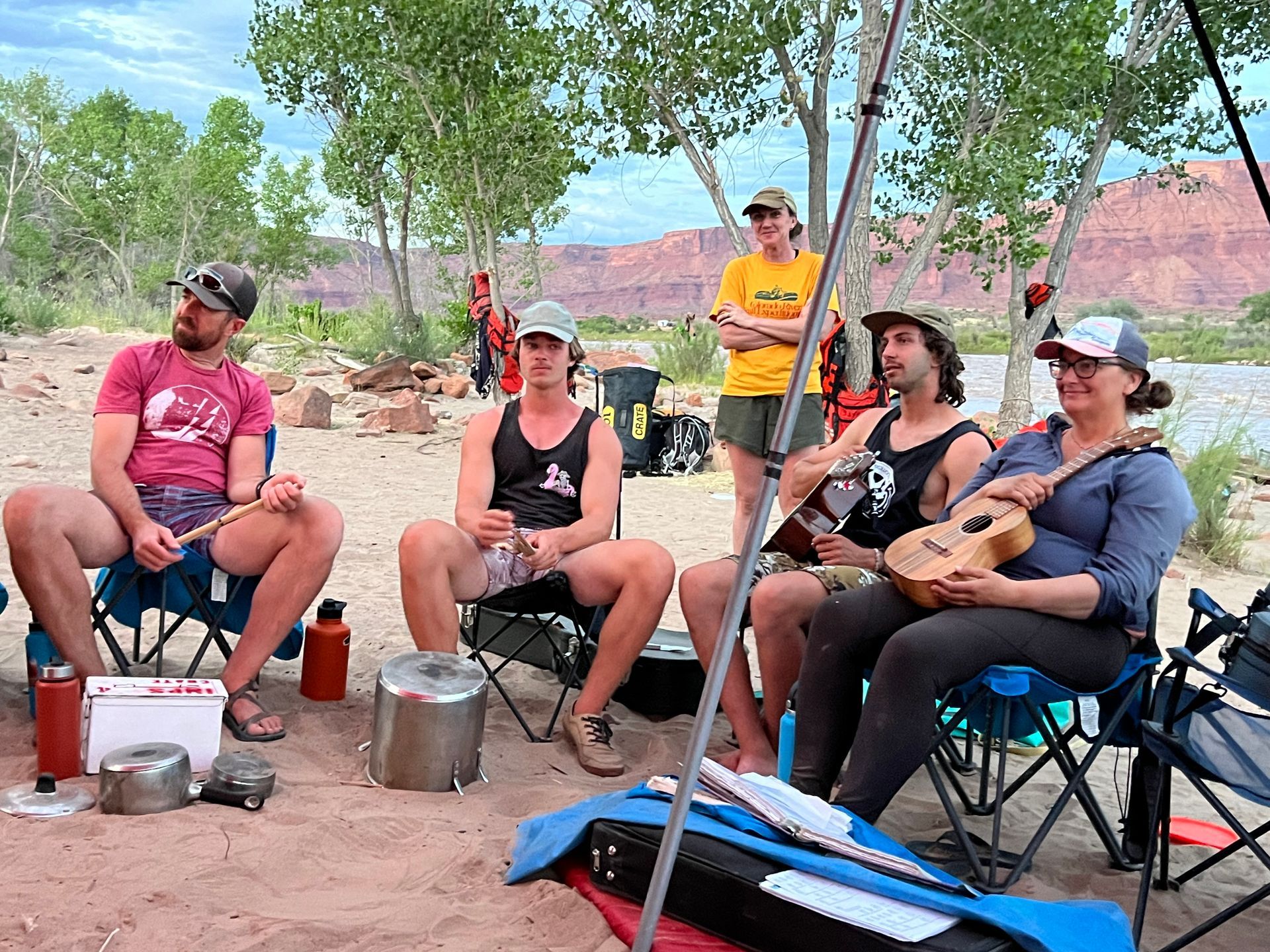 Group of people sitting in chairs outdoors by a river, some playing ukulele.