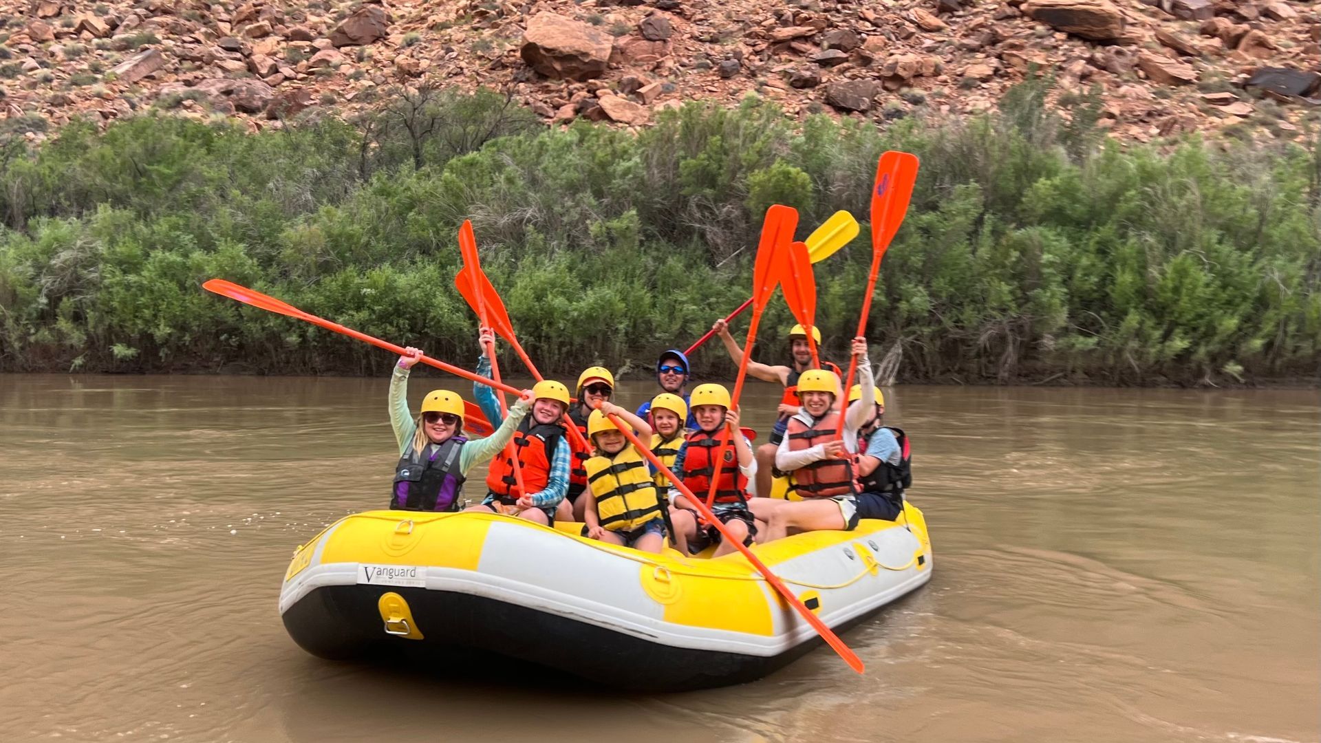 Group of people in yellow raft, raising paddles on river. Brown canyon backdrop.