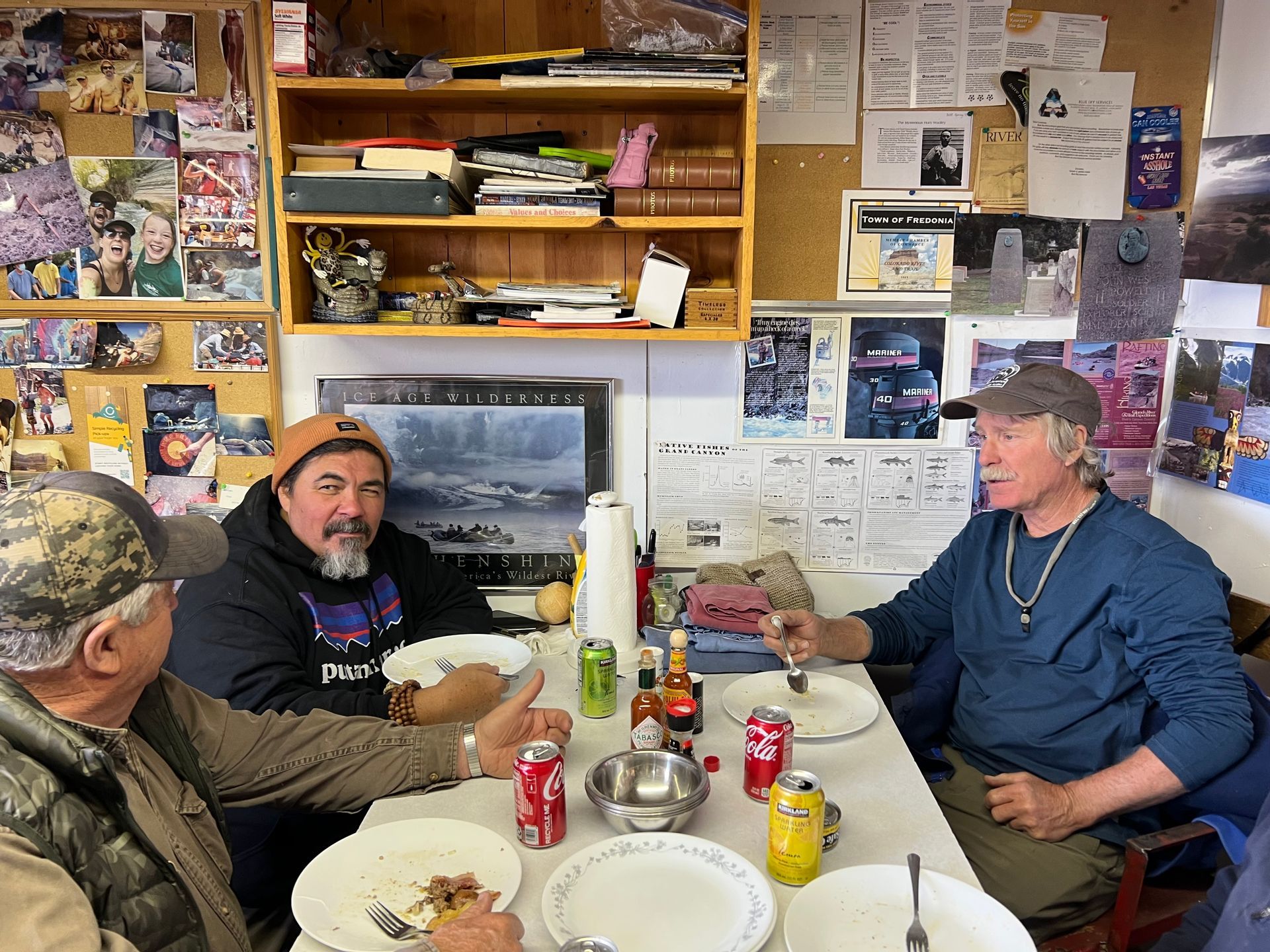 Three men seated at a table, eating. Interior shot with walls covered in items.