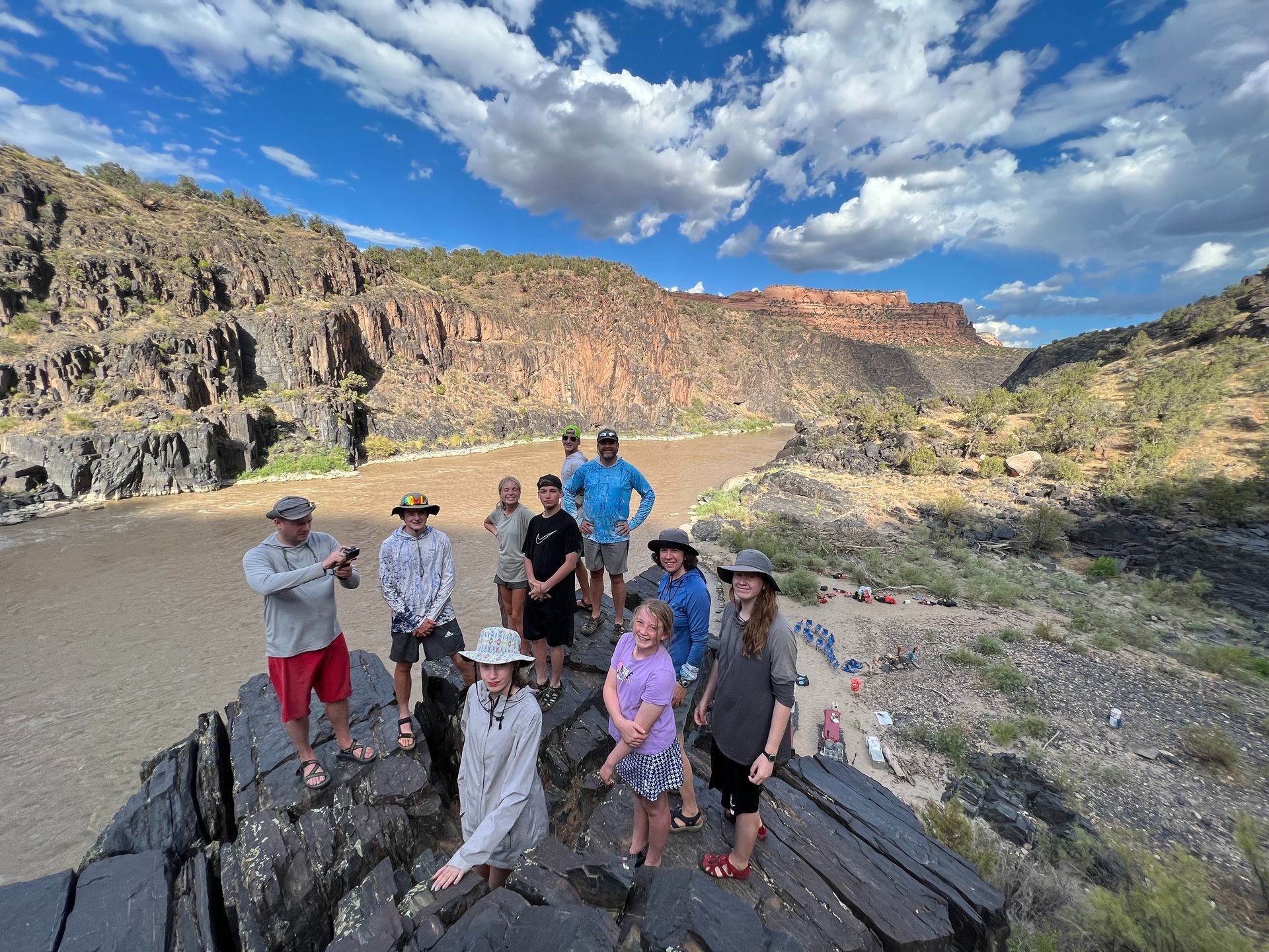 River with green foliage in front of a large red rock cliff under a blue sky.
