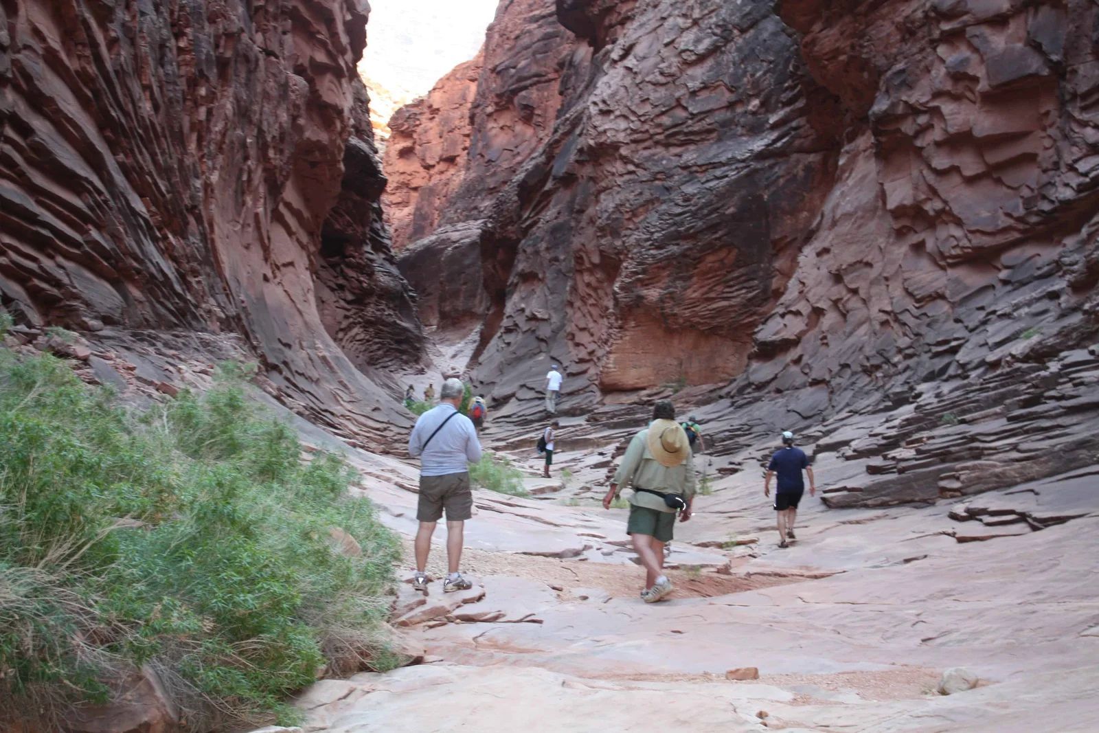 Hikers walk through a narrow, red-rock canyon.  Sunlight illuminates the path.