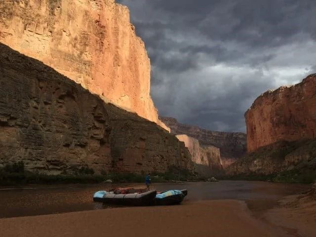 Rafts on muddy riverbank, canyon walls in sunlight and shadow, dramatic cloudy sky.