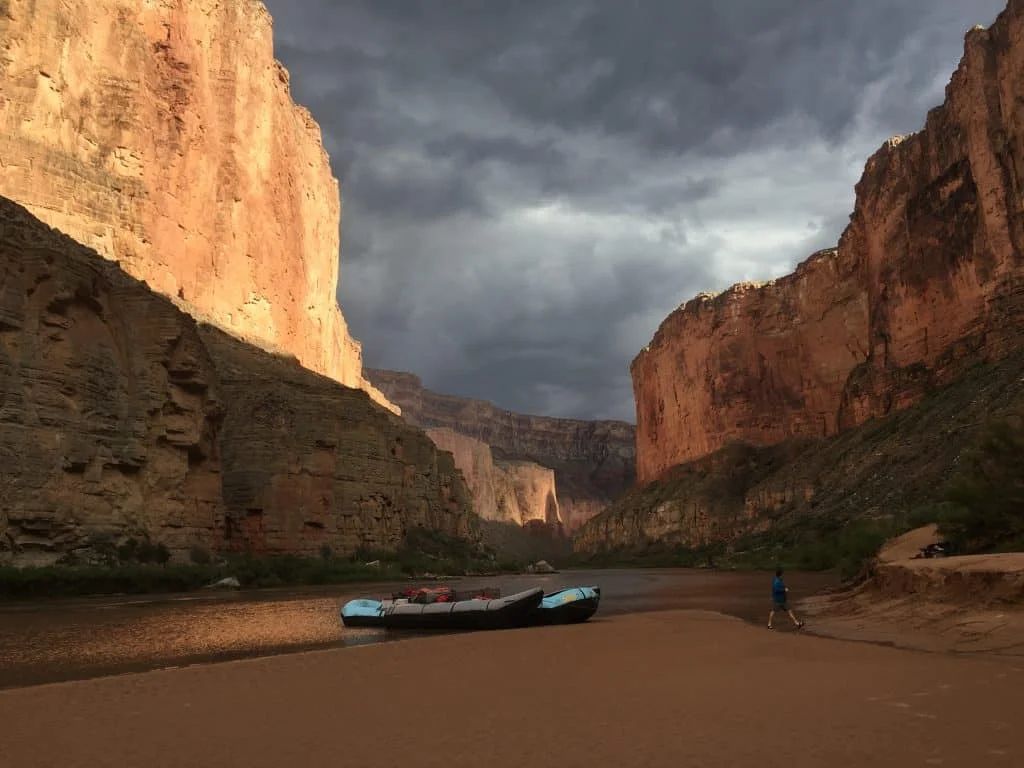 Raft on sandy riverbank in canyon, under dramatic, cloudy sky.