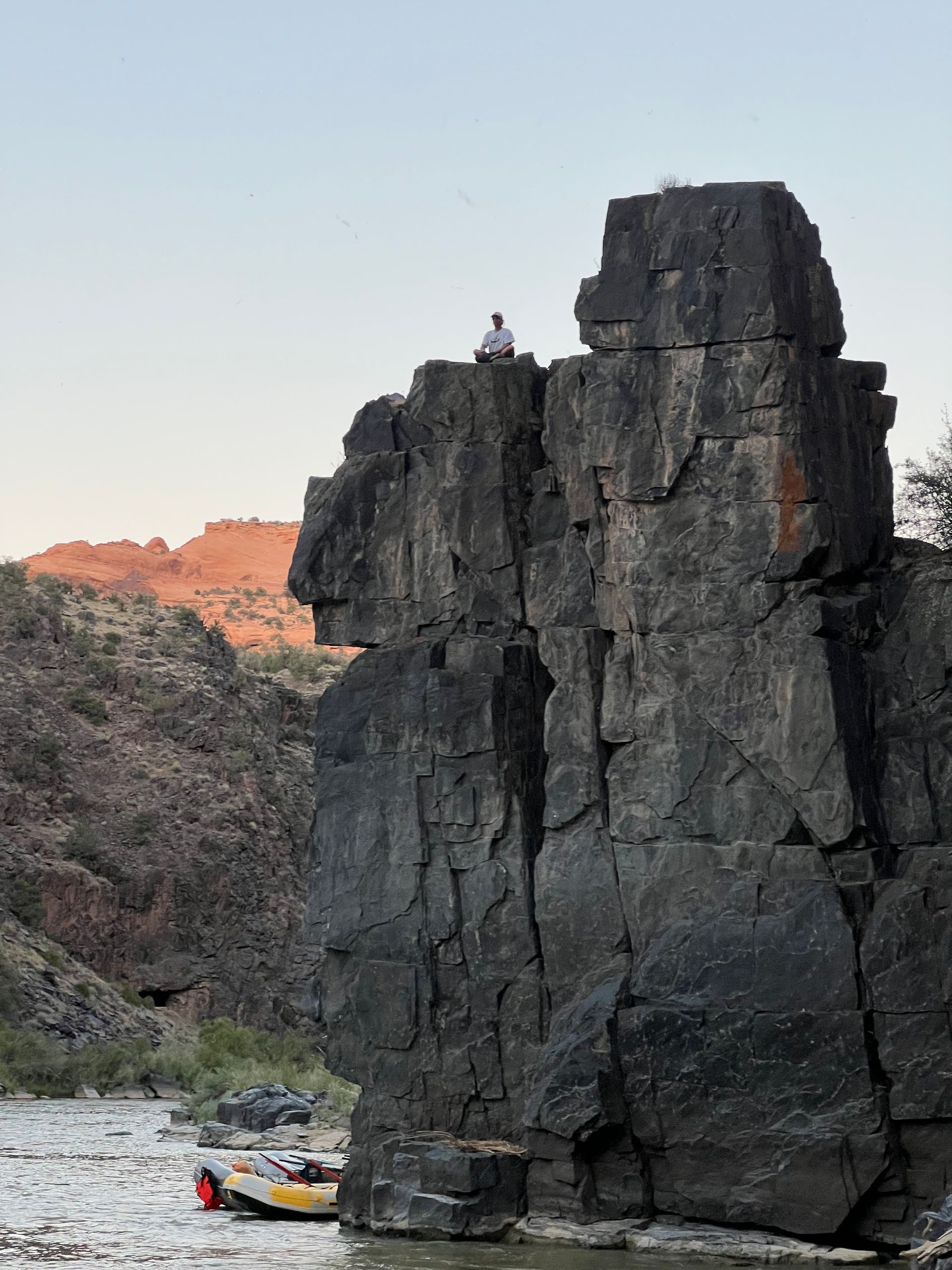 Person sits atop a large rock formation overlooking a river; a raft rests in the water.