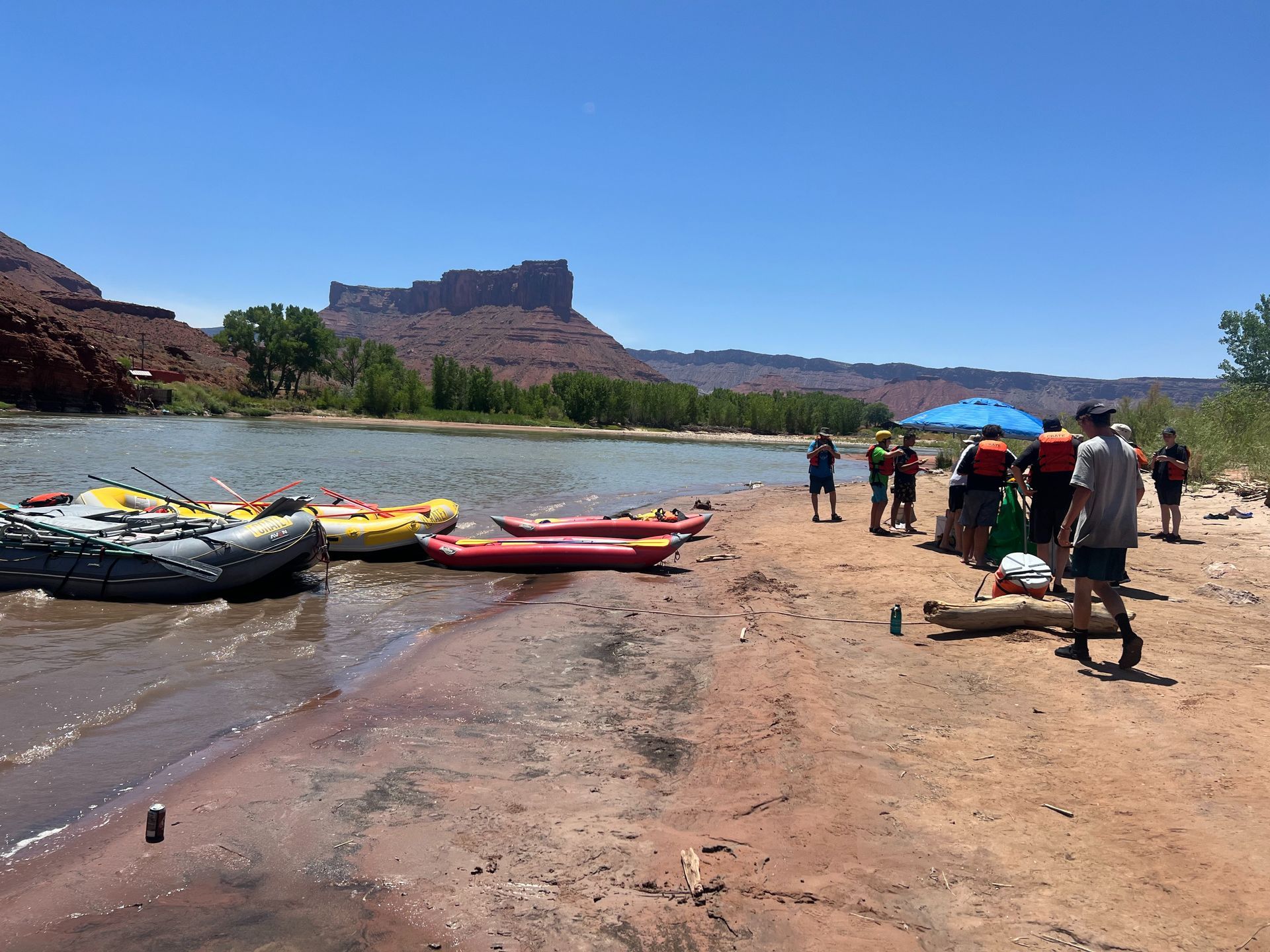 Kayaks and a raft on a sandy shore; people gather near a river and red rock formations.