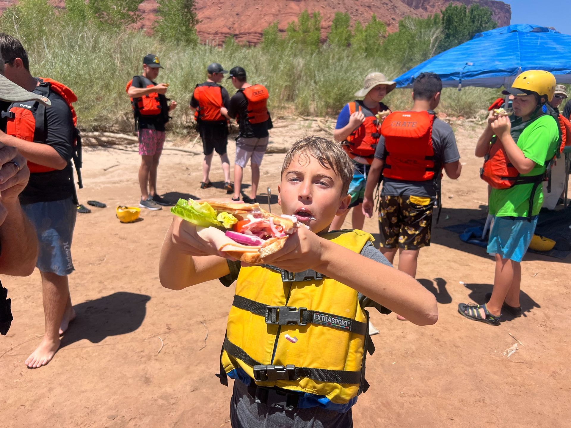 Boy eating a sandwich, wearing a life jacket, outdoors near a river with others.