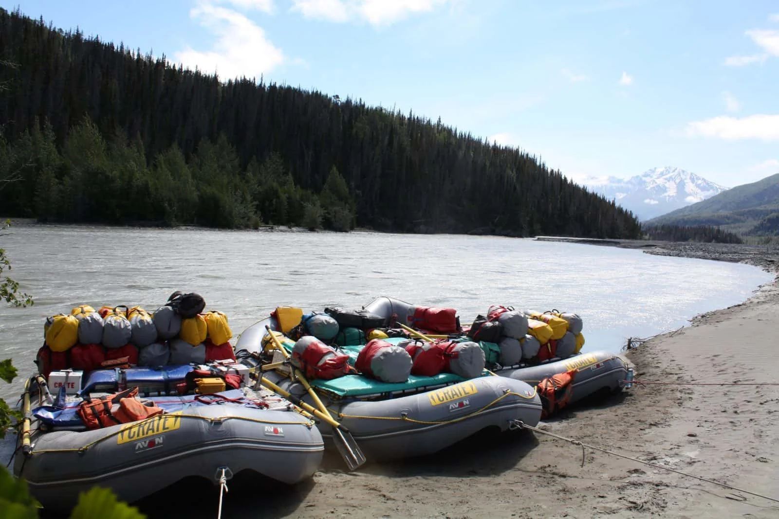 Three rafts loaded with gear on a sandy riverbank, with a forest and mountain in the background.