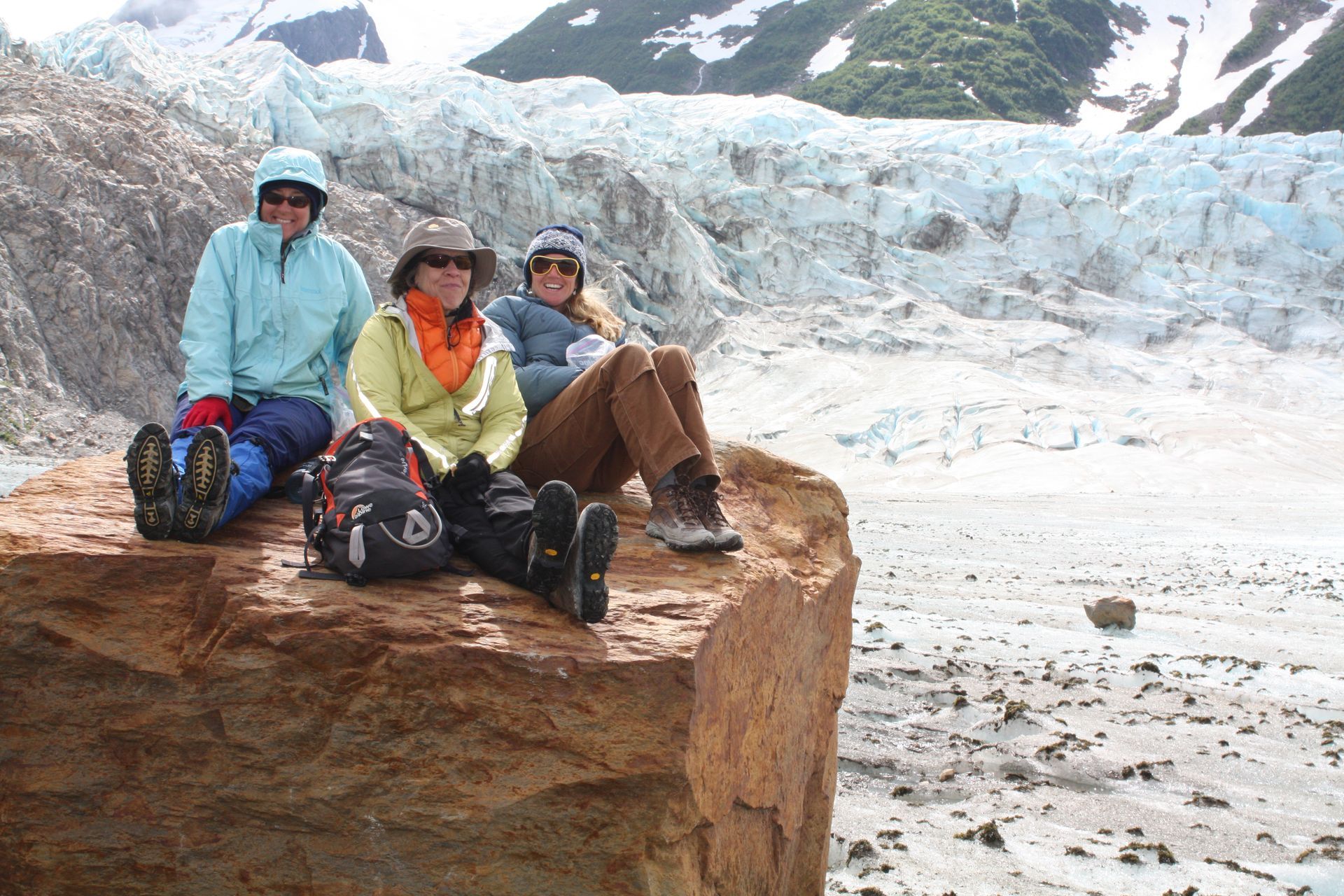 Three people sit on a rock, overlooking a glacier. One wears light blue, the others yellow and brown jackets.