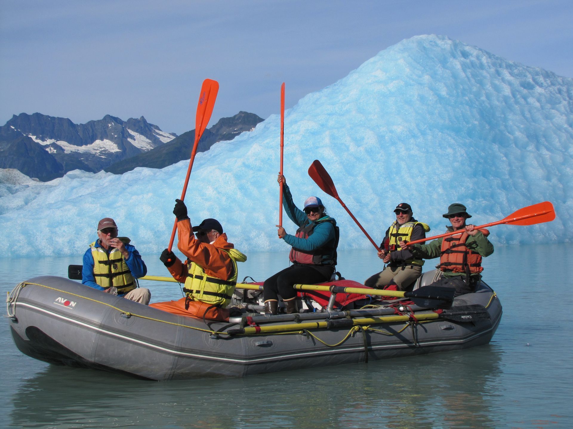 People rafting on a river, raising paddles, in front of a blue iceberg.