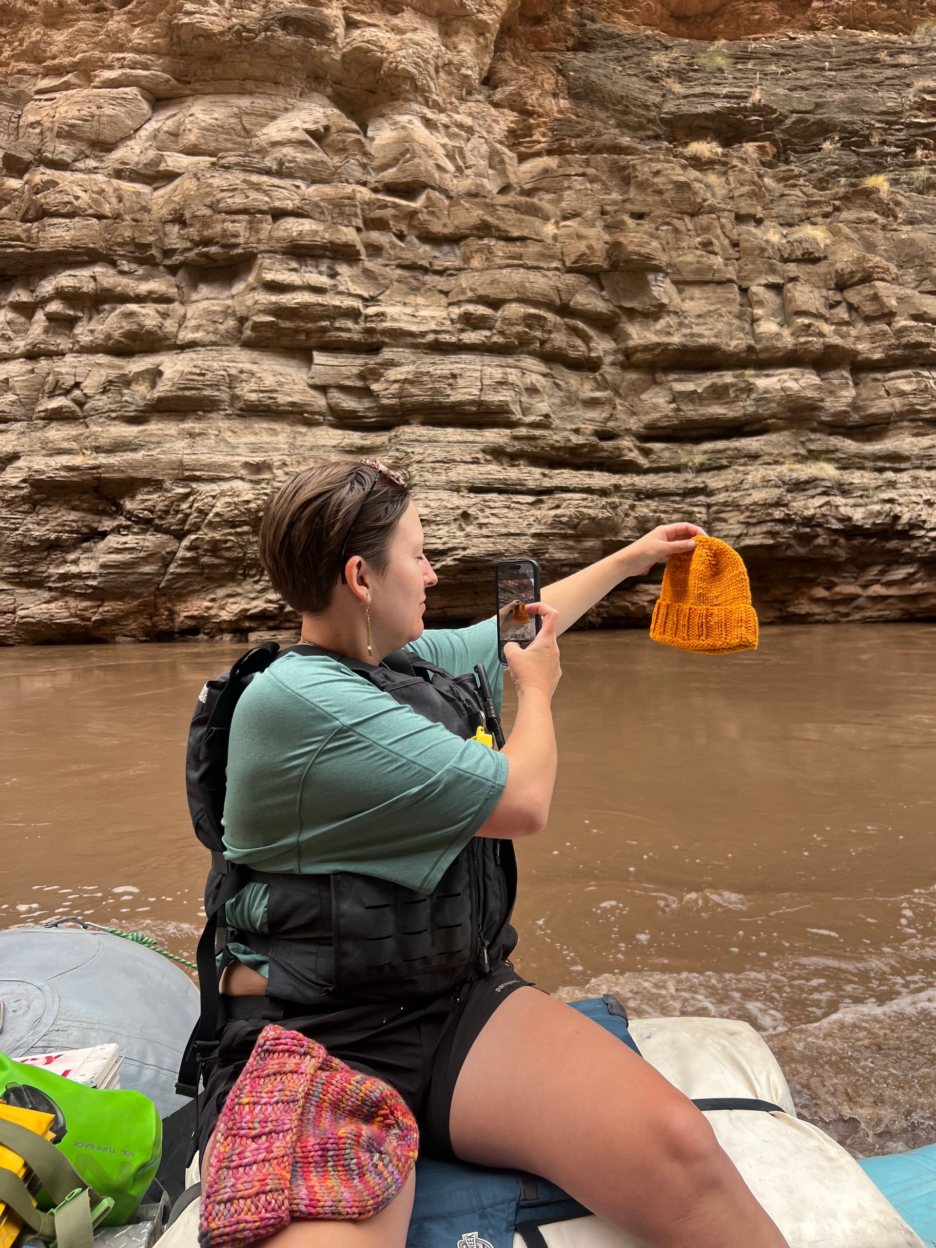 Woman on raft, holds up orange hat, taking photo. River and rock wall background.