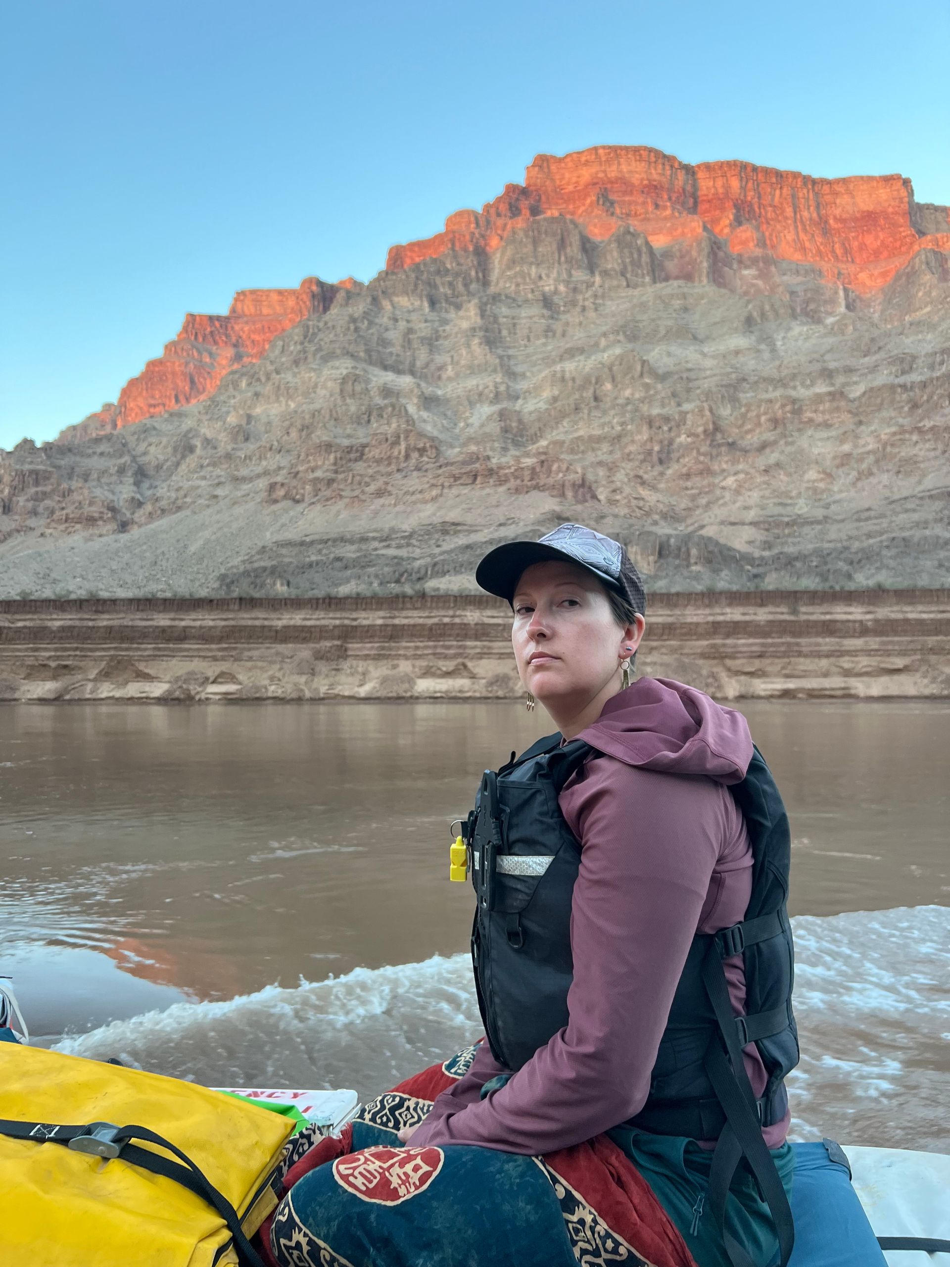 Woman in life vest on a boat, facing forward. Mountain in the background at sunset, river below.