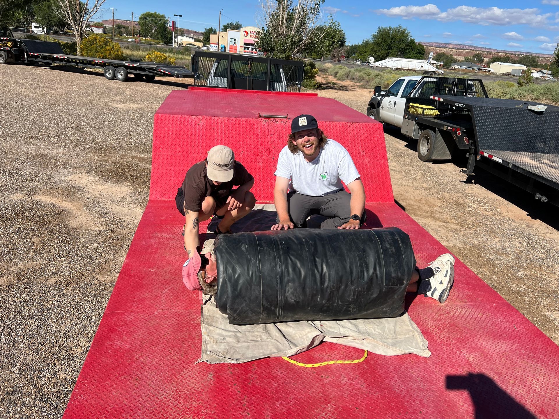 Two men on a red surface, unrolling a large black object. Outdoors, sunny day.