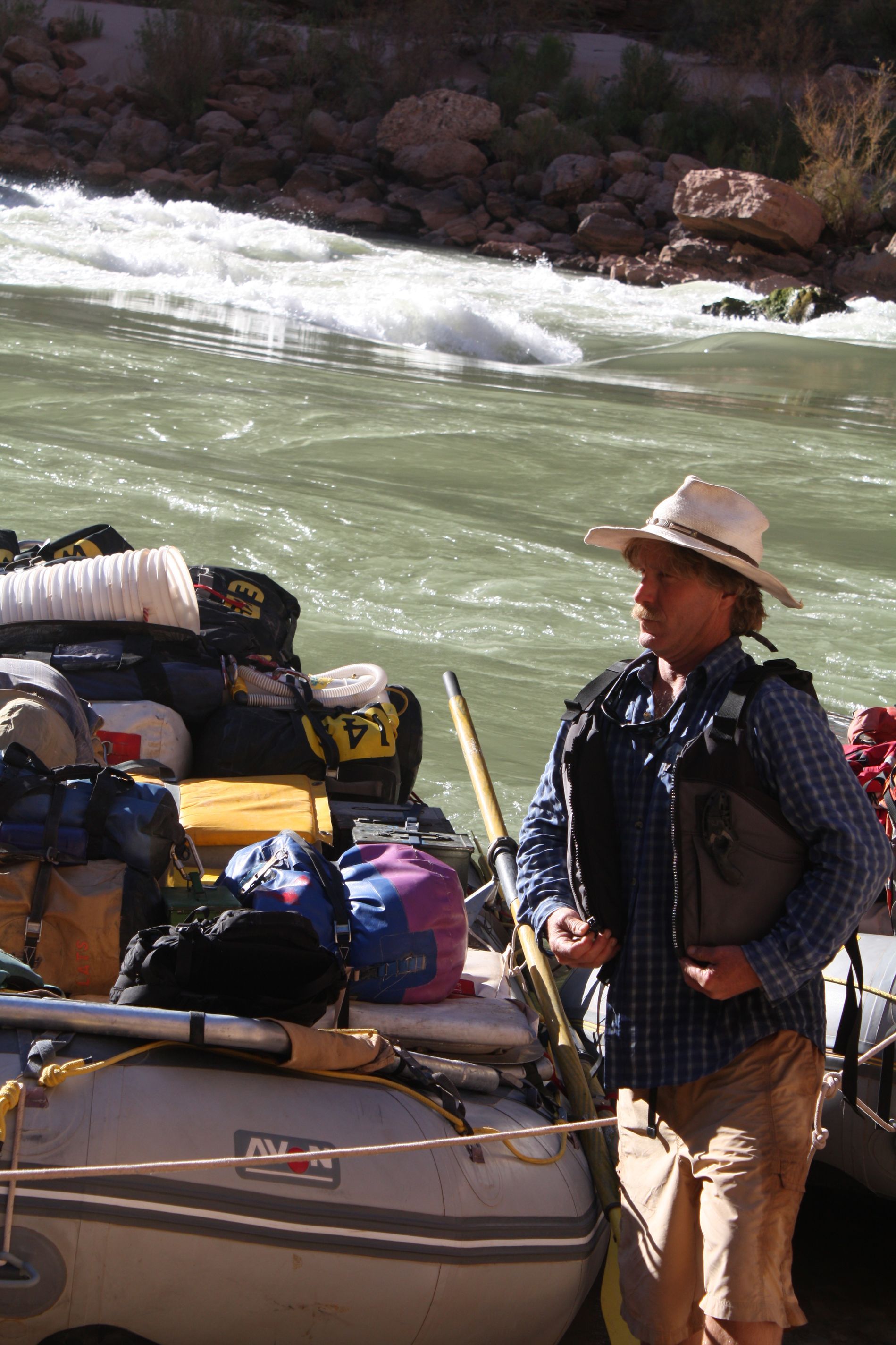 Person wearing a hat and life vest stands by a raft loaded with gear on a river.