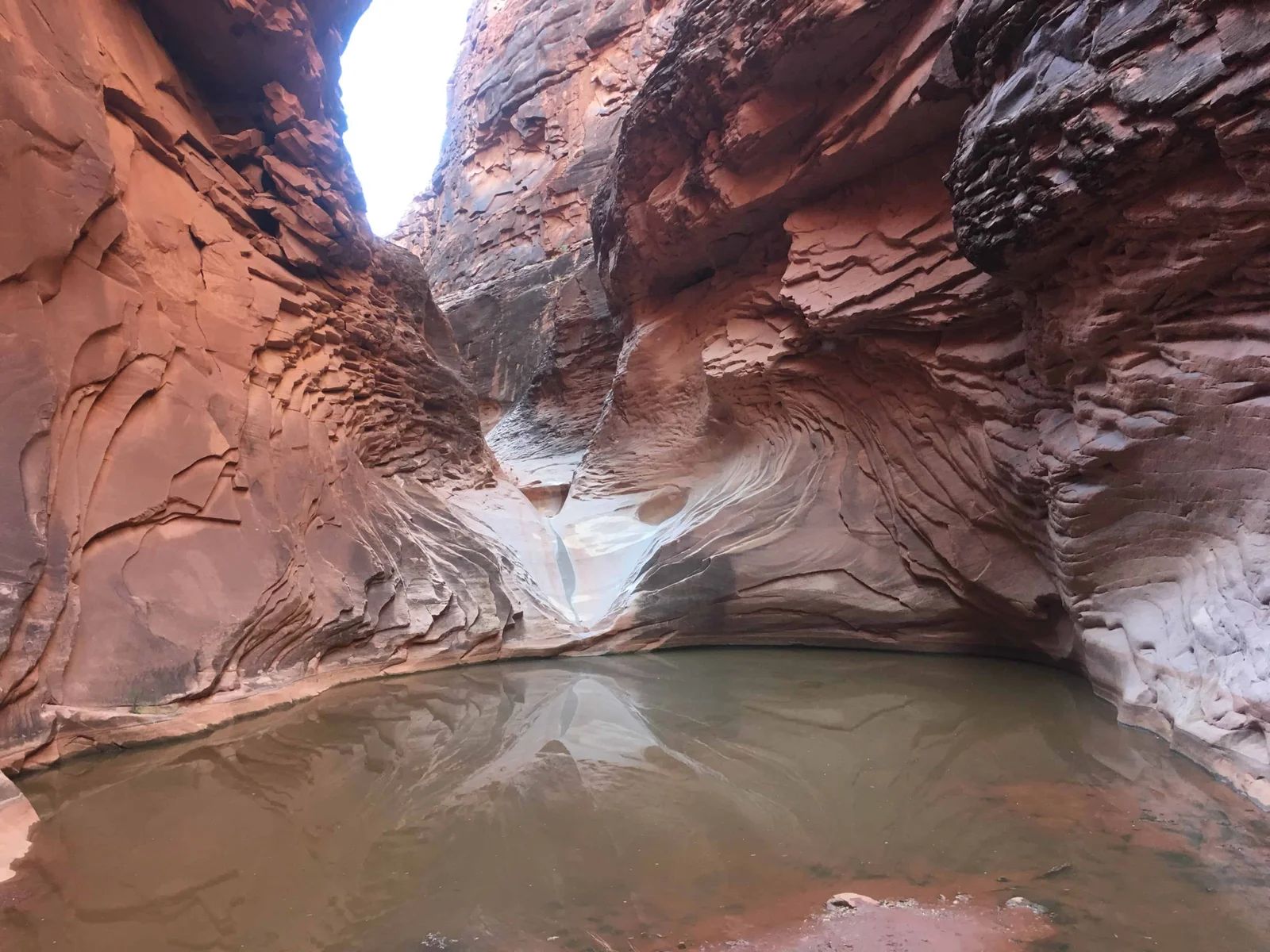 Narrow canyon with water-filled pool reflecting the sandstone walls and a glimpse of the sky.
