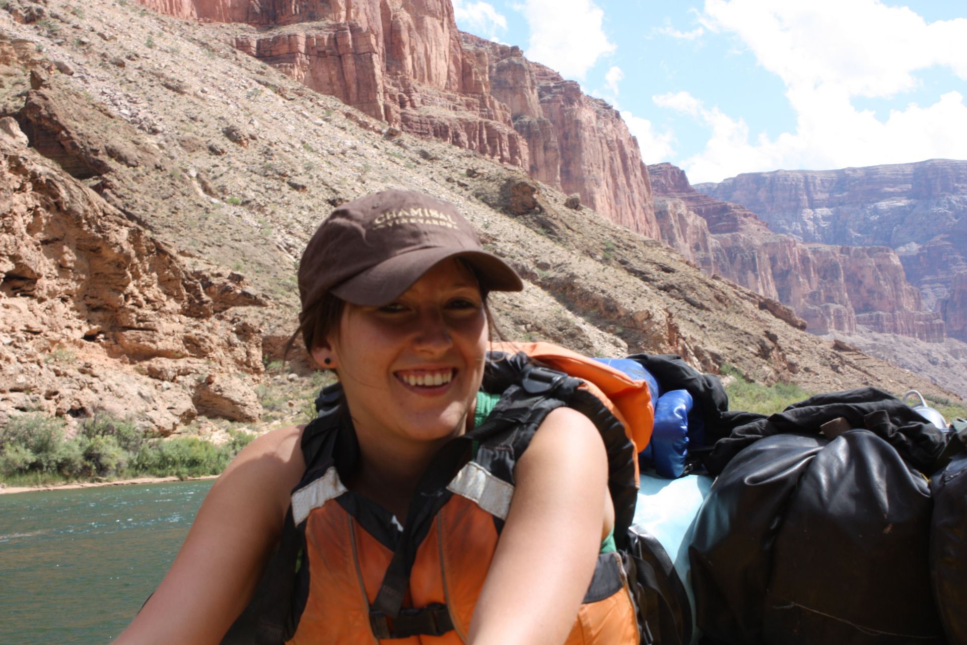 Woman smiling in a raft wearing a life vest and hat, with canyon walls in the background.