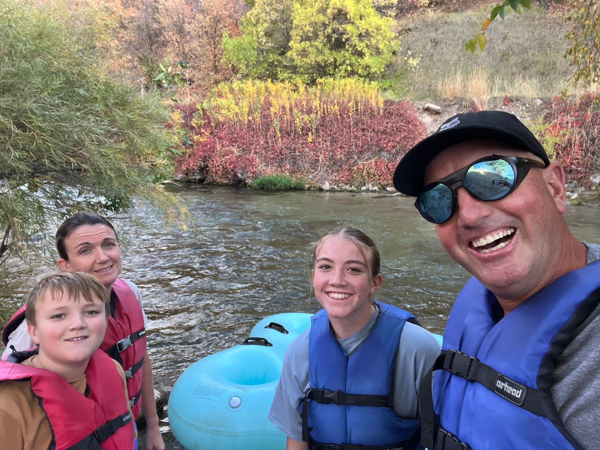 Family smiles for a selfie while river tubing, fall foliage backdrop.