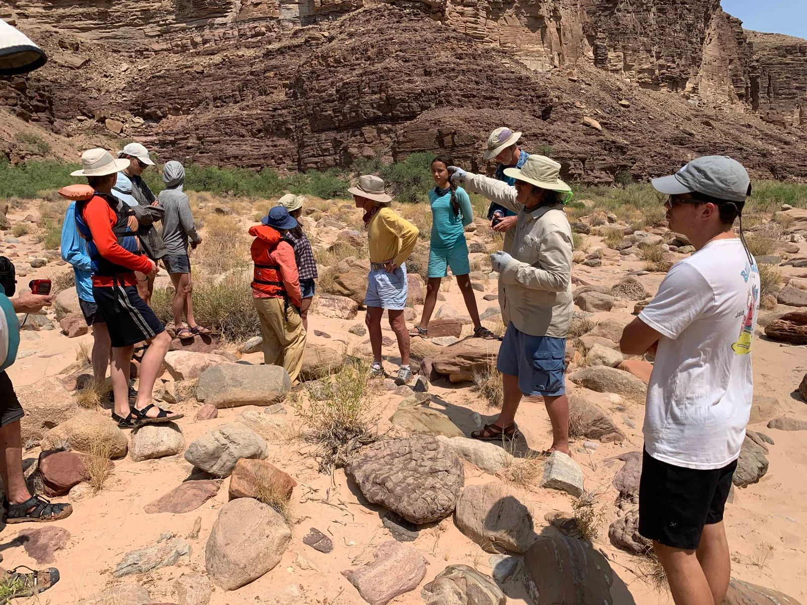 Group of people on a rocky terrain near a canyon. Some wear hats, looking at something pointed to.