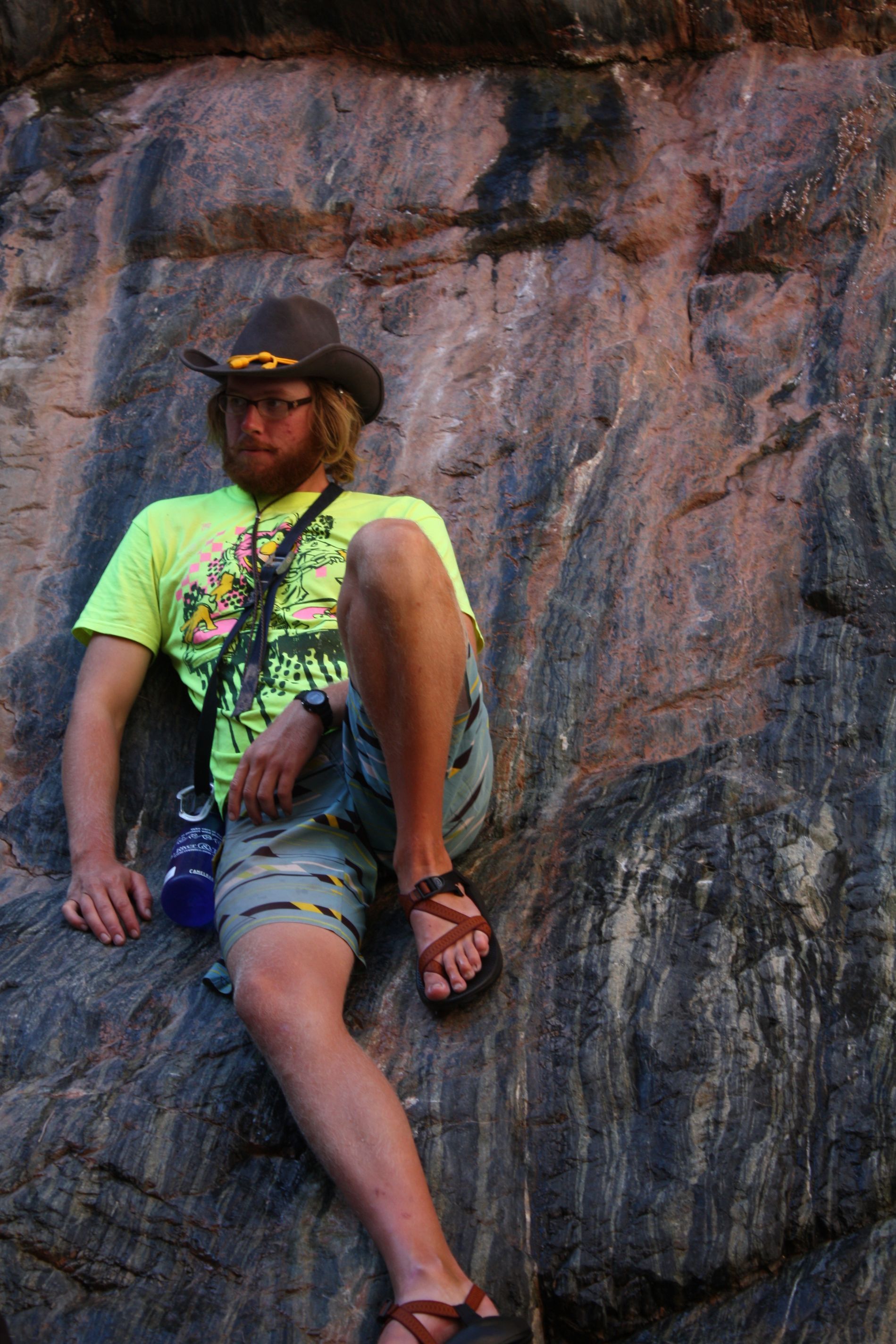 Man wearing cowboy hat and sandals leans against rock face.