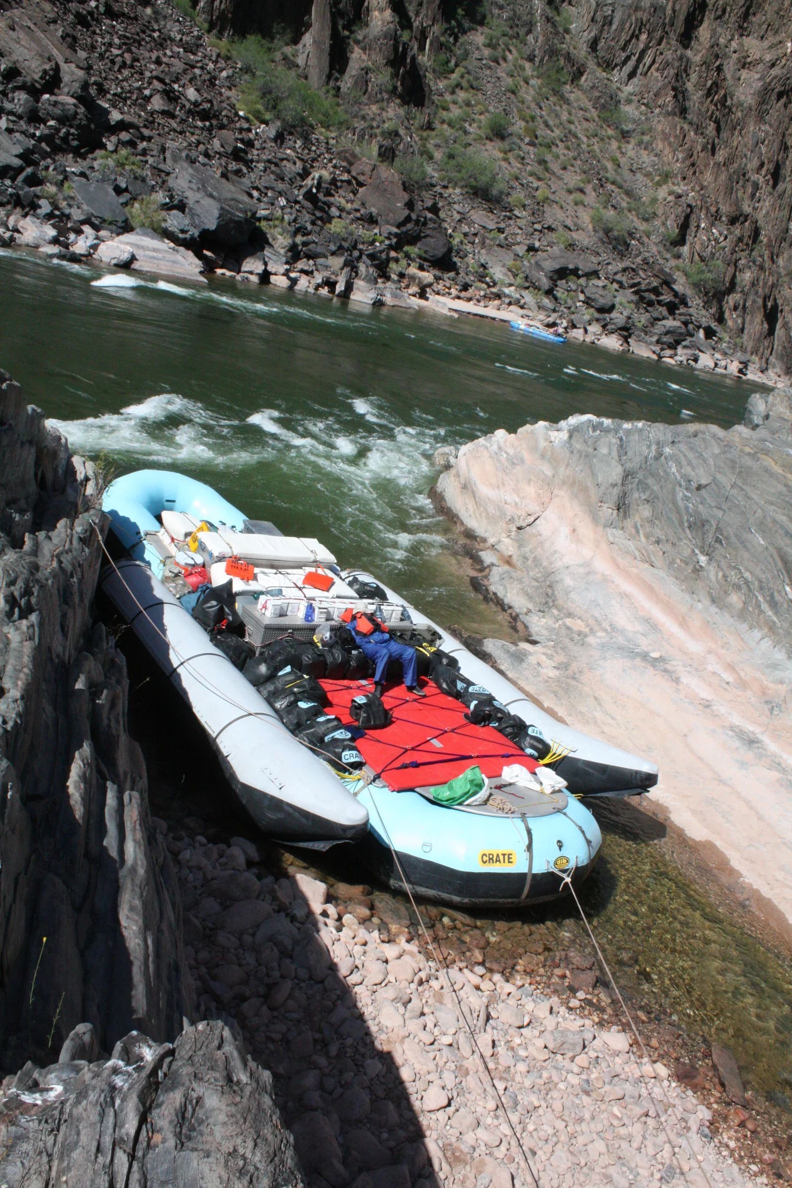 Raft pulled ashore next to a river with canyon walls. Blue and white raft with gear on board.