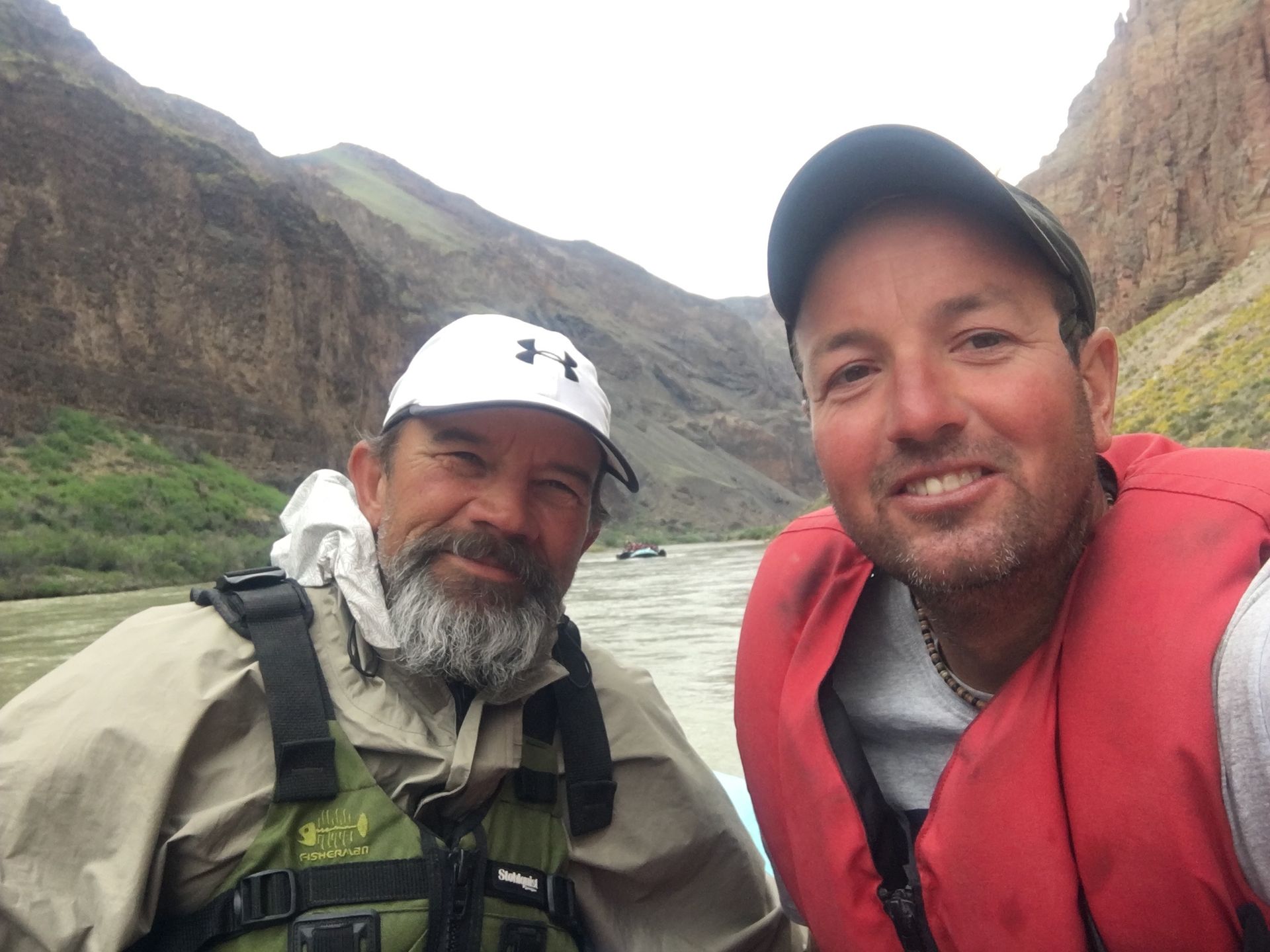 Two men on a river raft; one wearing a cap and jacket, the other a life vest; background of canyon walls.