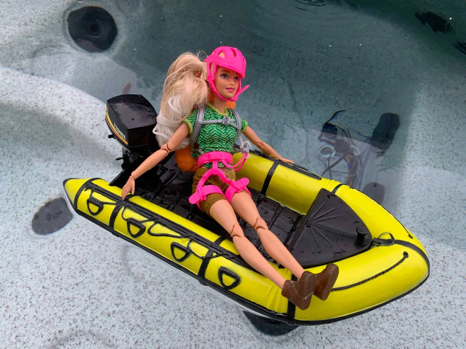 Barbie doll in yellow inflatable boat on water, wearing pink helmet and outdoor gear.